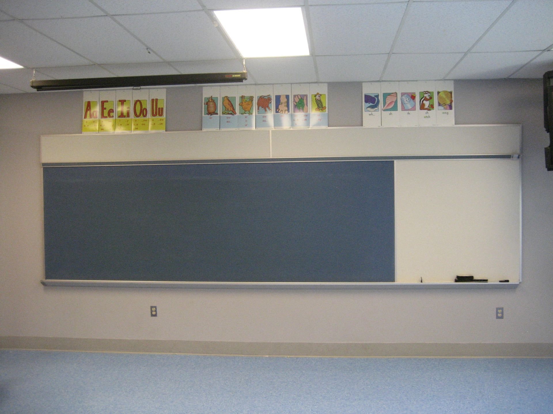 Empty classroom wall with a large blue chalkboard and whiteboard beneath a row of posters.