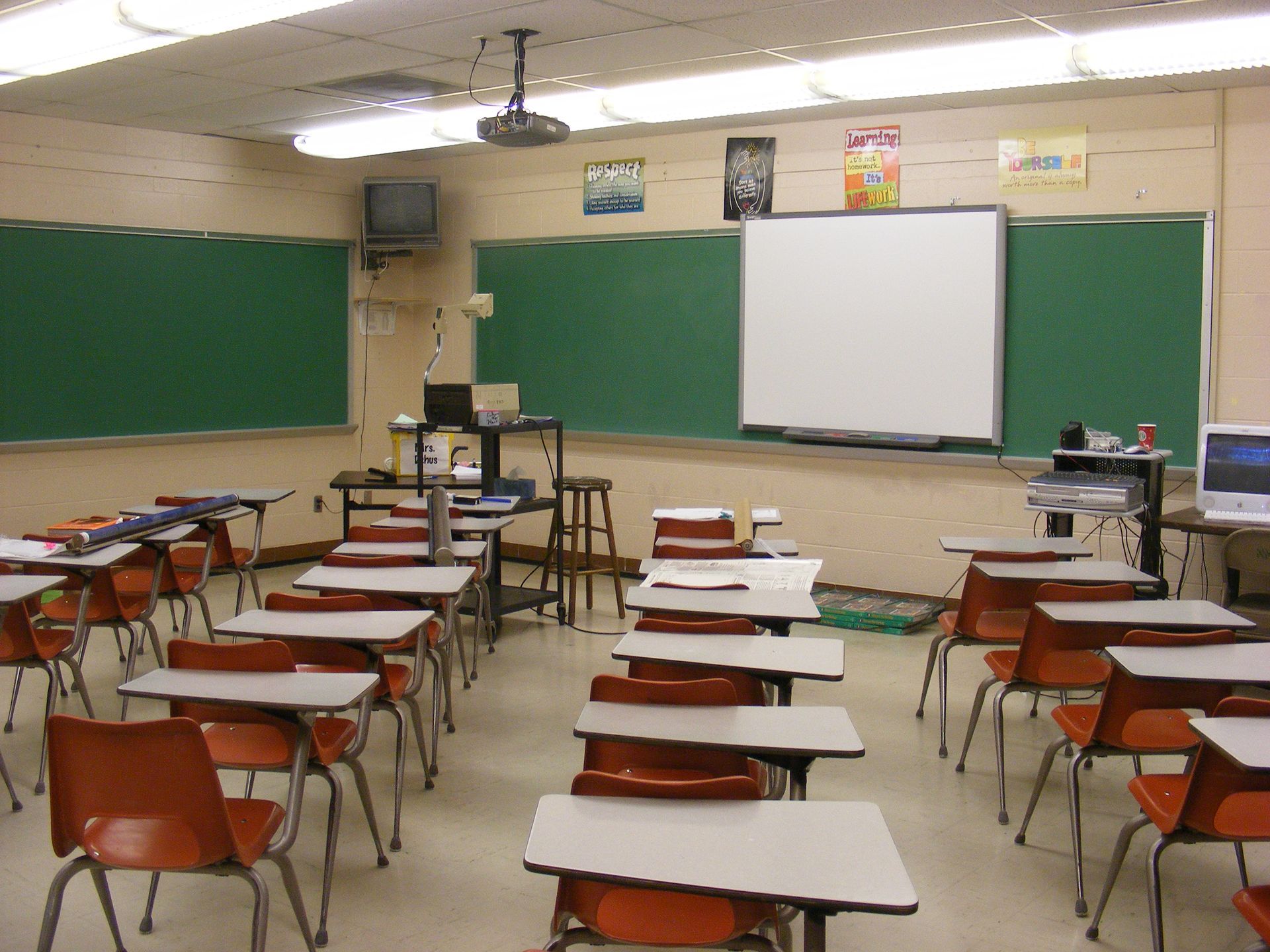Empty classroom with desks, green chalkboards, and a pull-down projector screen at the front.