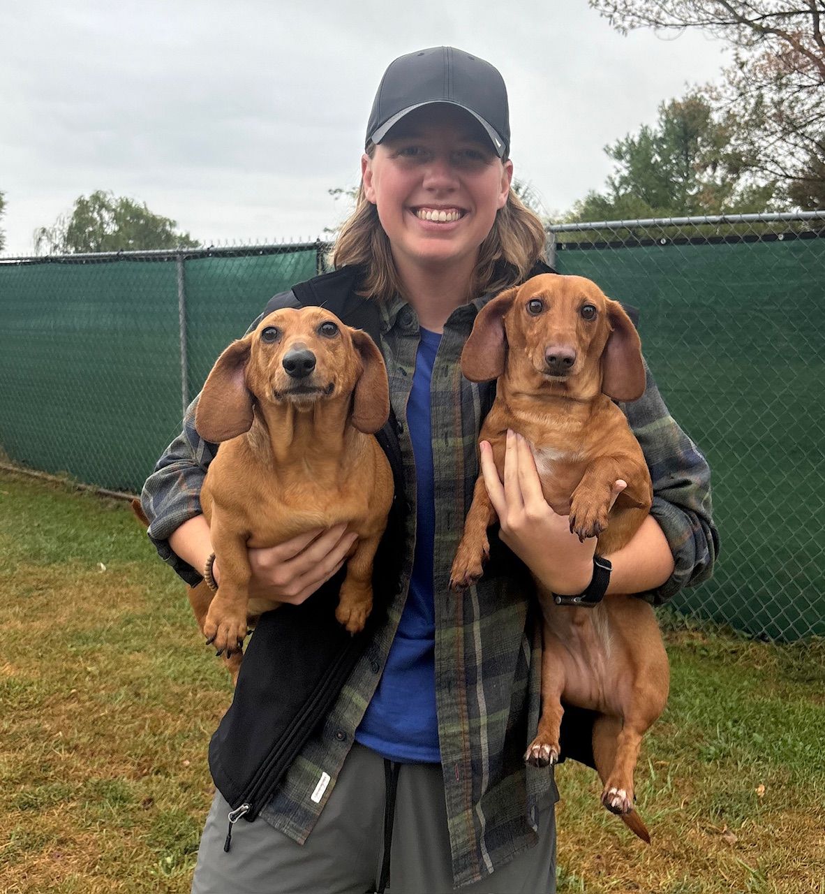 Woman holding two brown dachshunds outside.