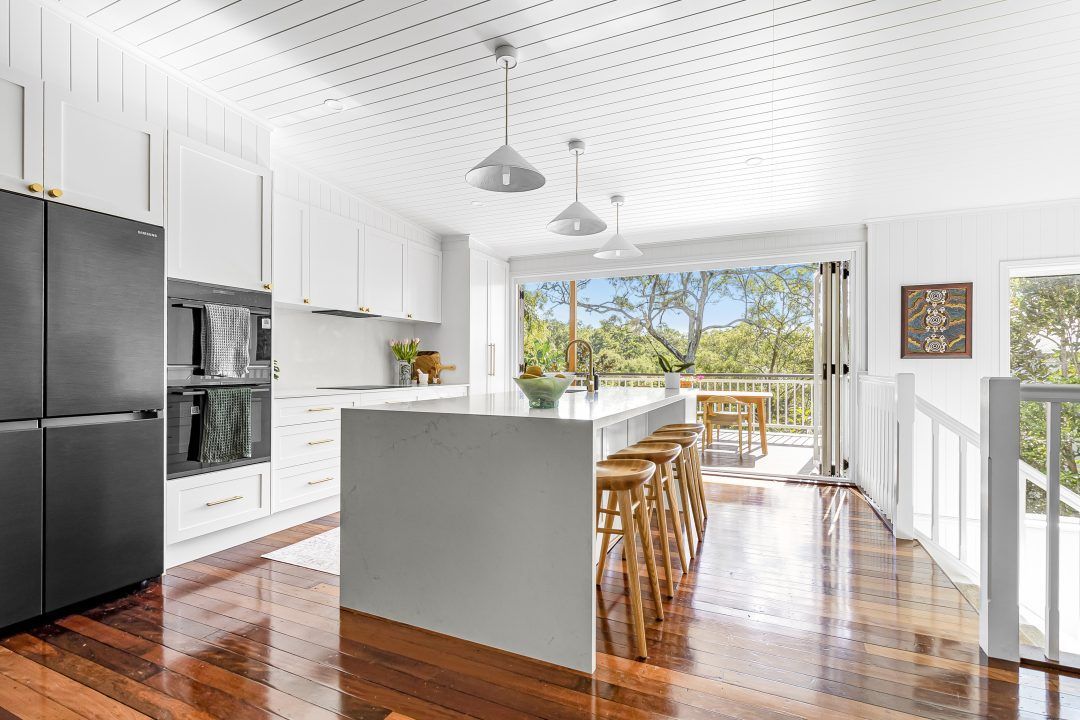 White kitchen with island, stools, and deck access. Dark refrigerator, wooden floors, and bright natural light.
