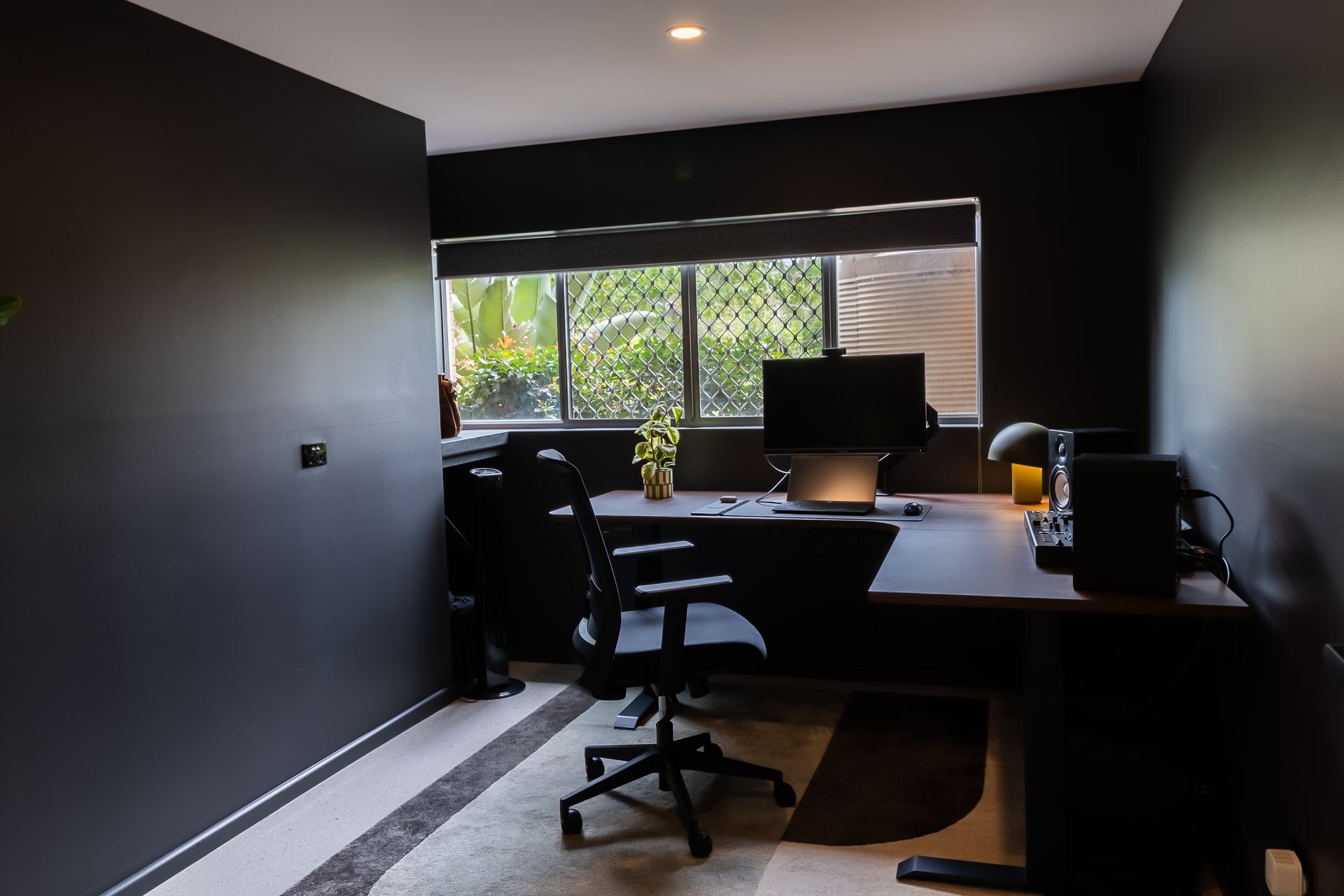 Black home office with desk, computer, chair, window, and greenery.