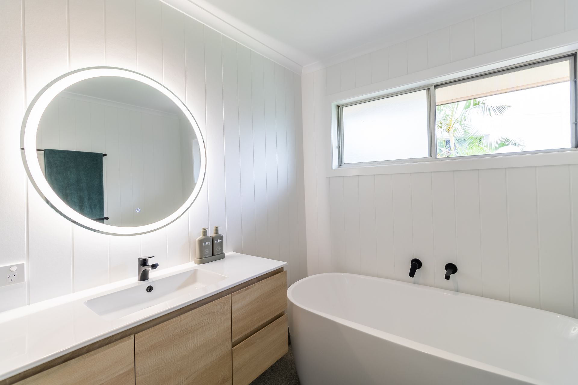 Modern white bathroom with wooden vanity, round backlit mirror, and freestanding tub.