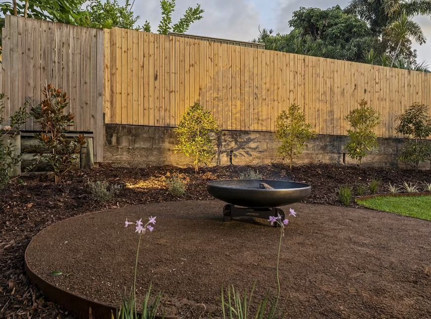 Round fire pit on gravel, surrounded by plants, in front of a wooden fence.