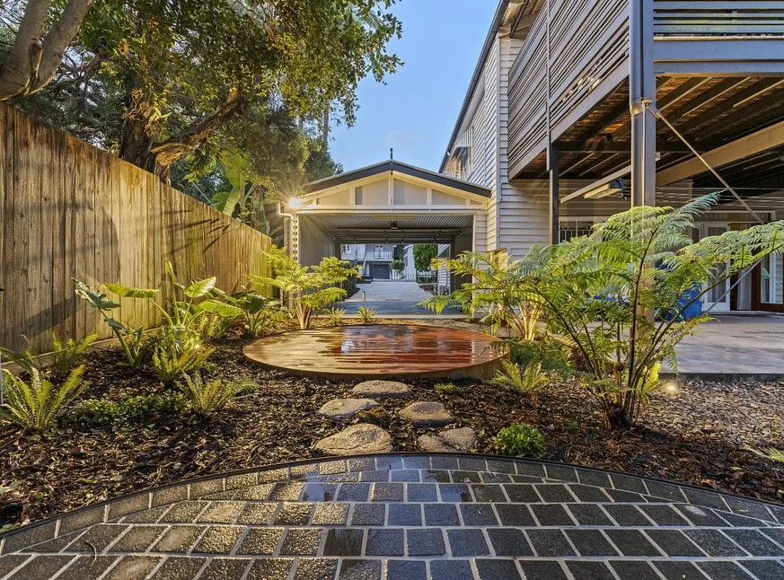 Well-lit garden path leads to a carport, flanked by greenery, a wooden fence, and a multi-level house.