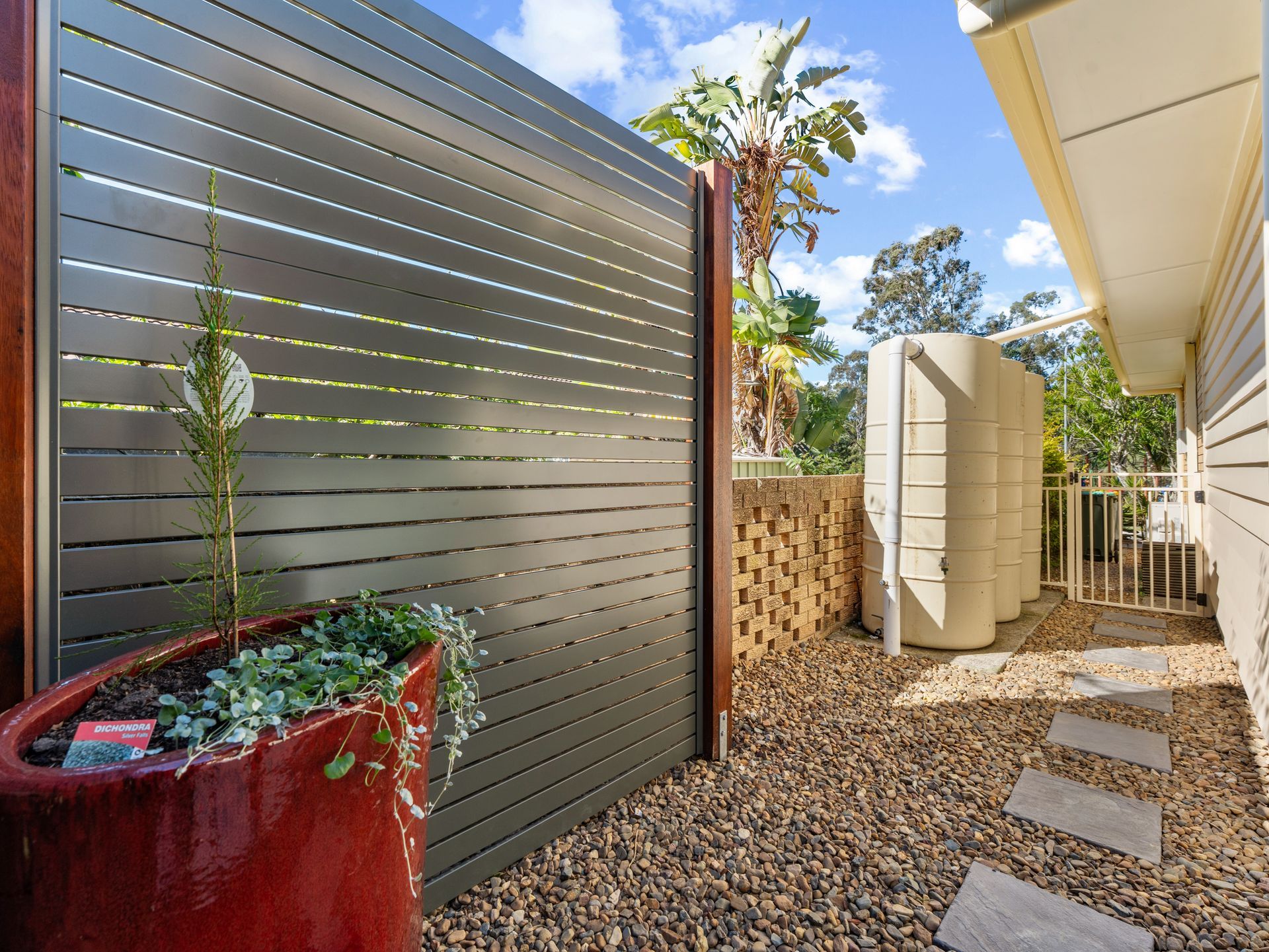 Dark slatted fence in a gravel yard. Red planter with plant, water tank, pavers, beige house and blue sky.