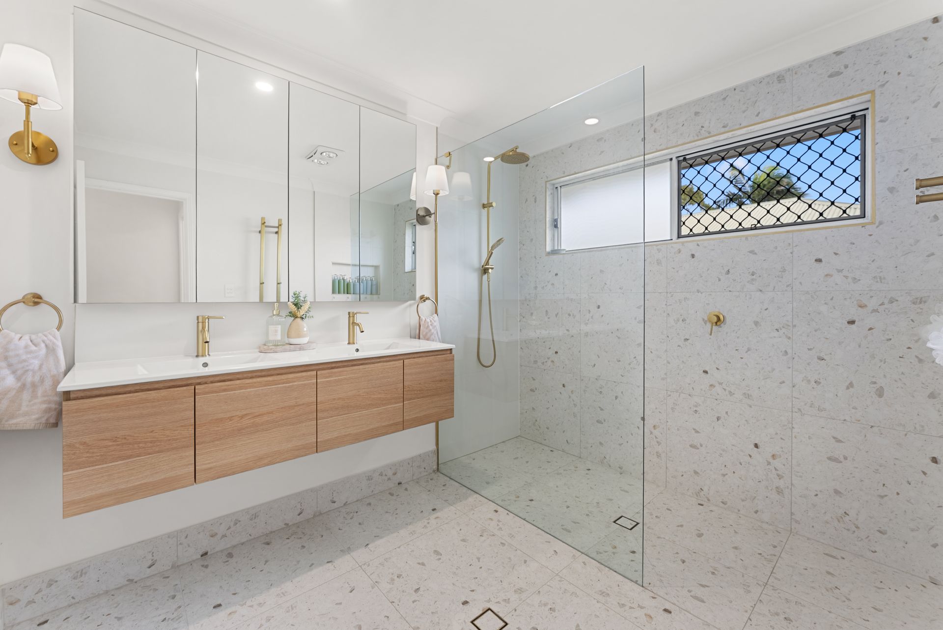 Modern white bathroom with wood vanity, large mirror, glass shower, and terrazzo tile flooring.