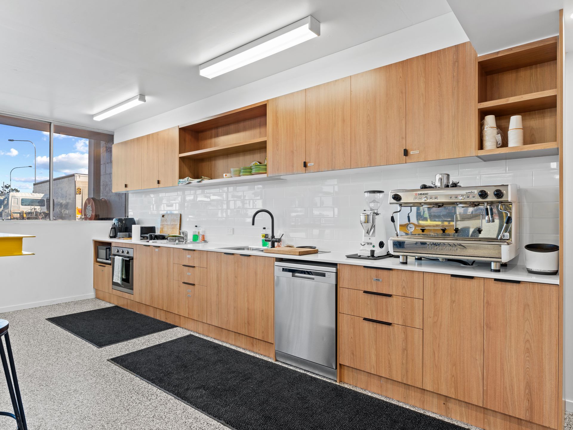 Modern office kitchen with light wood cabinets, stainless steel appliances, and black floor mats.