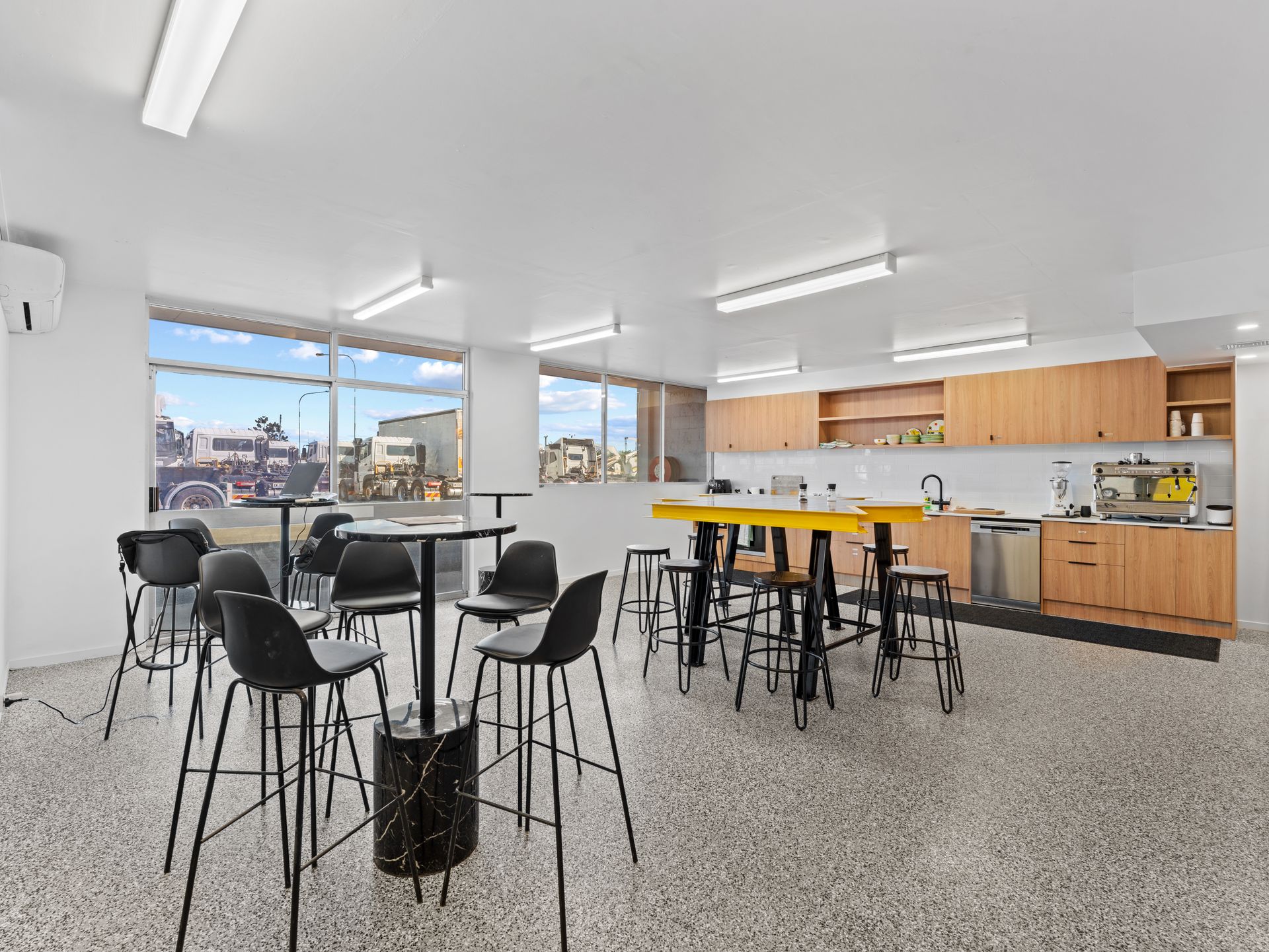 Modern office kitchen area with high-top tables, black chairs, and a yellow workstation; large windows.