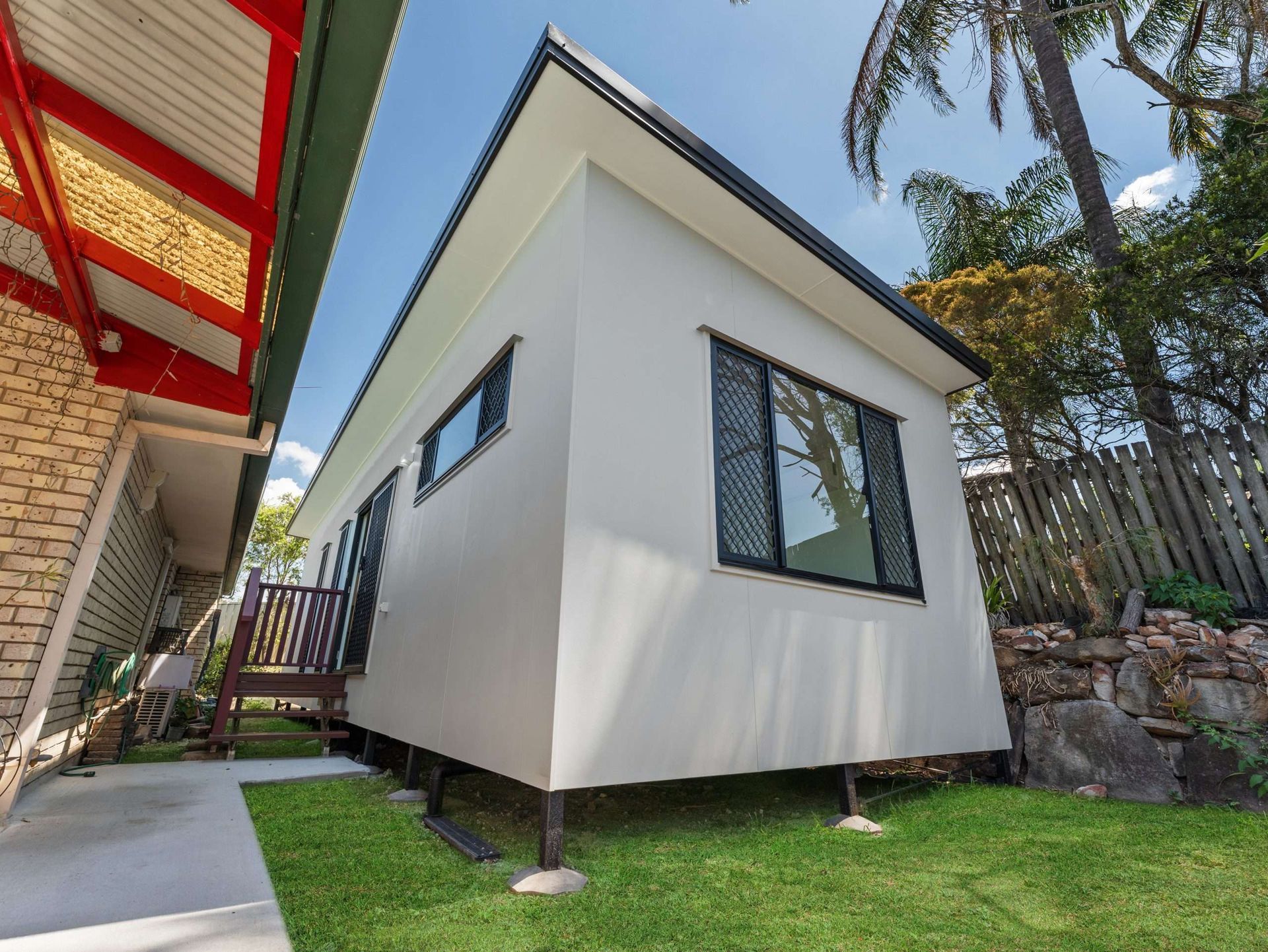 Small, modern, cream-colored home on stilts with black window shutters, set on green grass, between two other buildings.