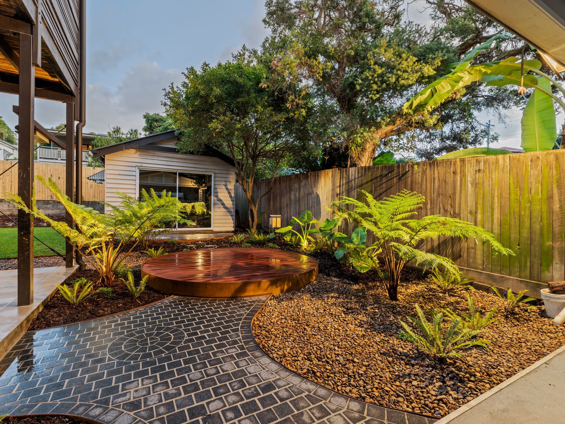Garden path of pavers leading to a rusted steel circular seating area, surrounded by plants and a small shed.