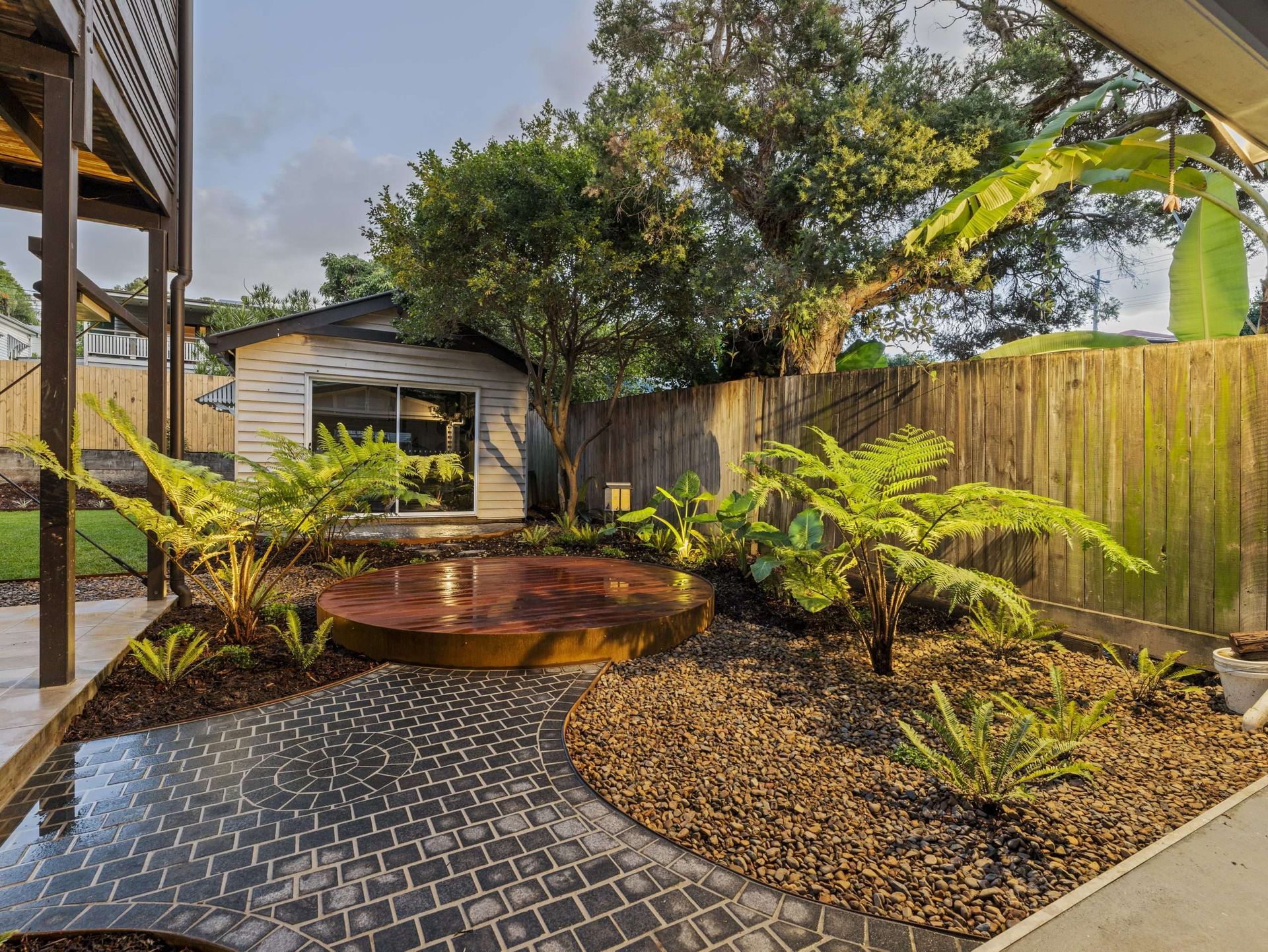 A backyard garden with a rusted metal water feature, ferns, and a winding stone path, with a shed in the background.