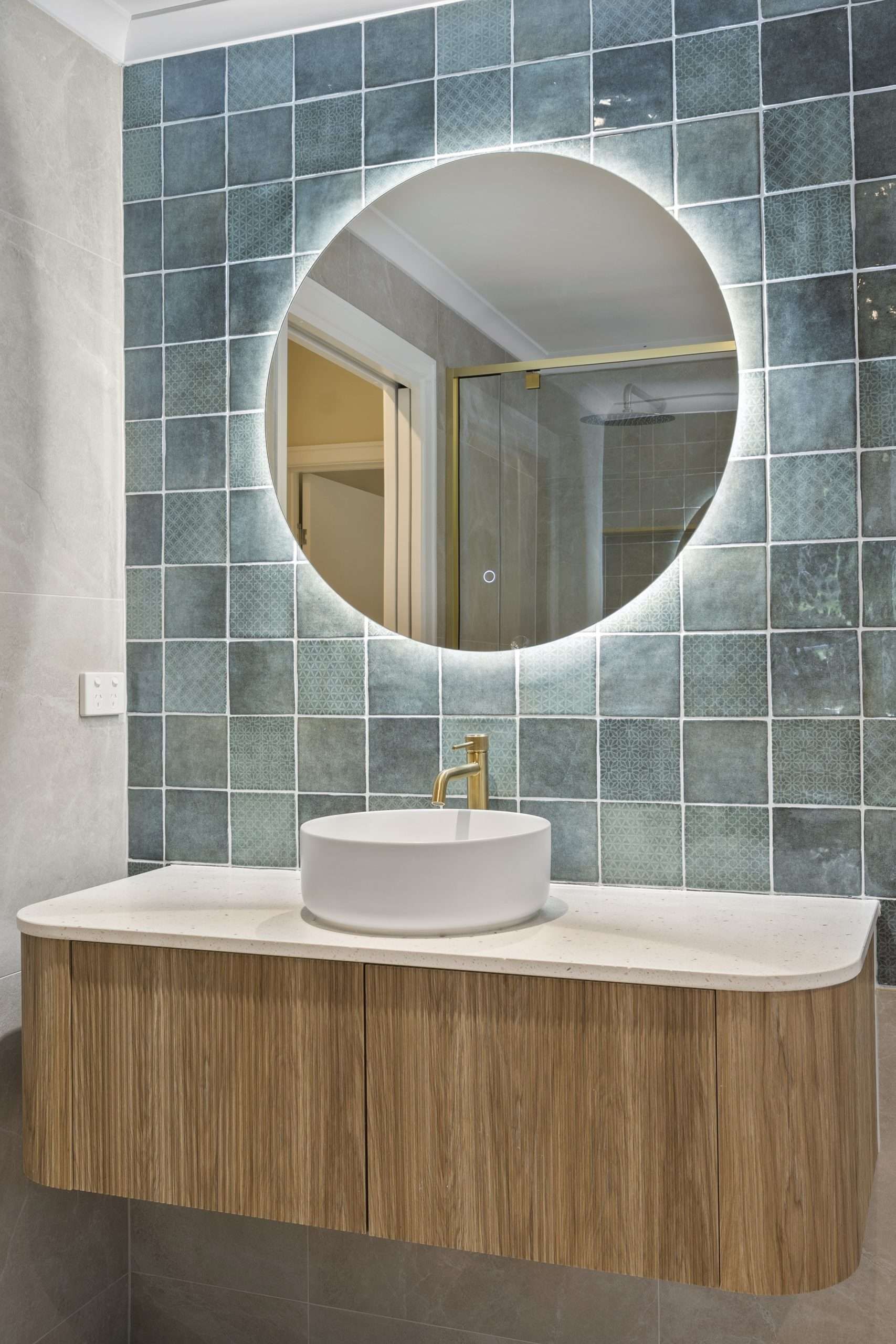 Bathroom with teal tiled wall, round mirror, wooden vanity, and white sink.