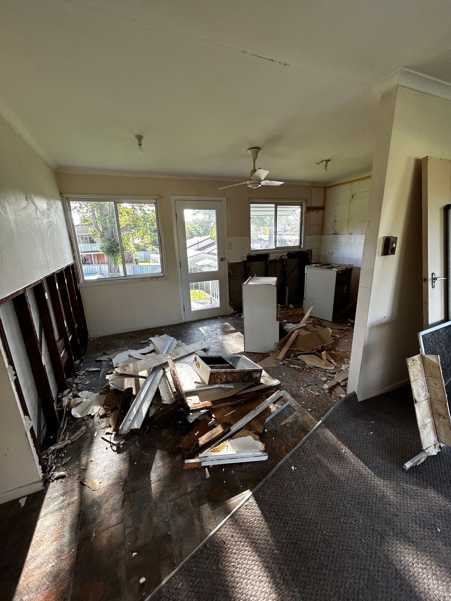 Interior of a demolished room; debris on the floor, exposed walls, doorway to outside, window, and ceiling fan.
