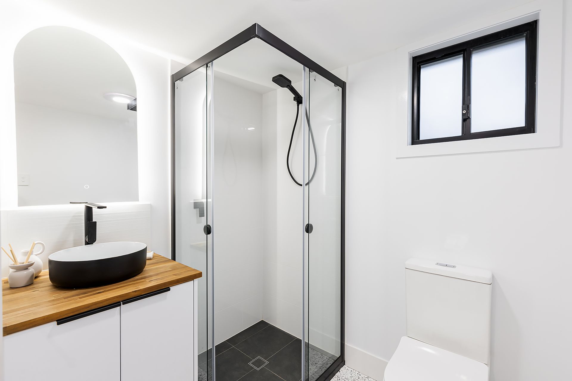 Modern bathroom: glass shower, black trim, white vanity with wooden top, black vessel sink, toilet, and window.