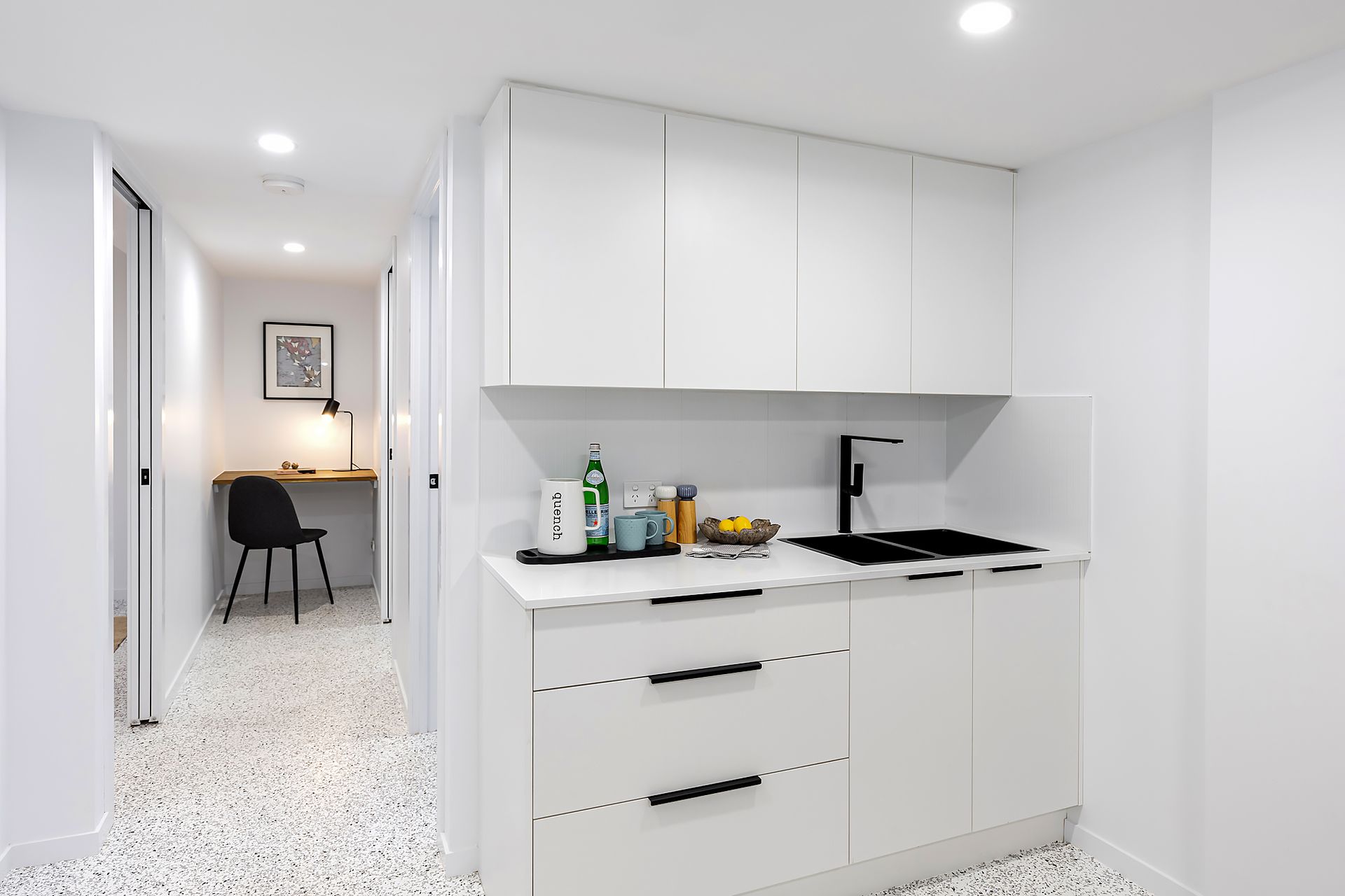 White kitchenette in a hallway with a desk and chair visible down the hall.