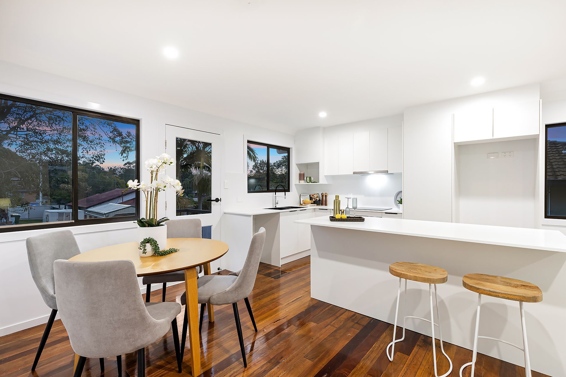Open-plan kitchen and dining area with wood floors, white cabinets, and gray chairs; windows overlook a tree-filled landscape.