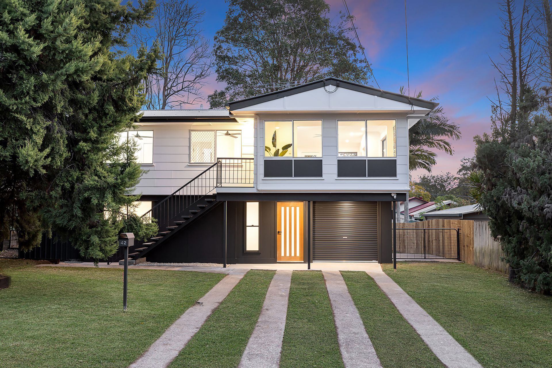Two-story white and black house with a driveway leading to a garage. Green lawn and trees surround.