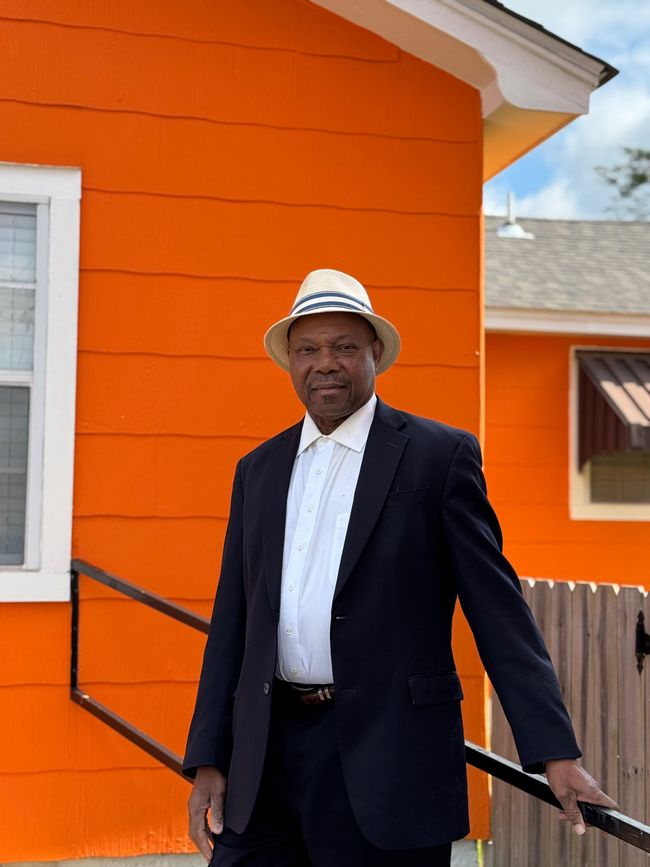 Man in a suit and hat stands in front of an orange building, smiling slightly.