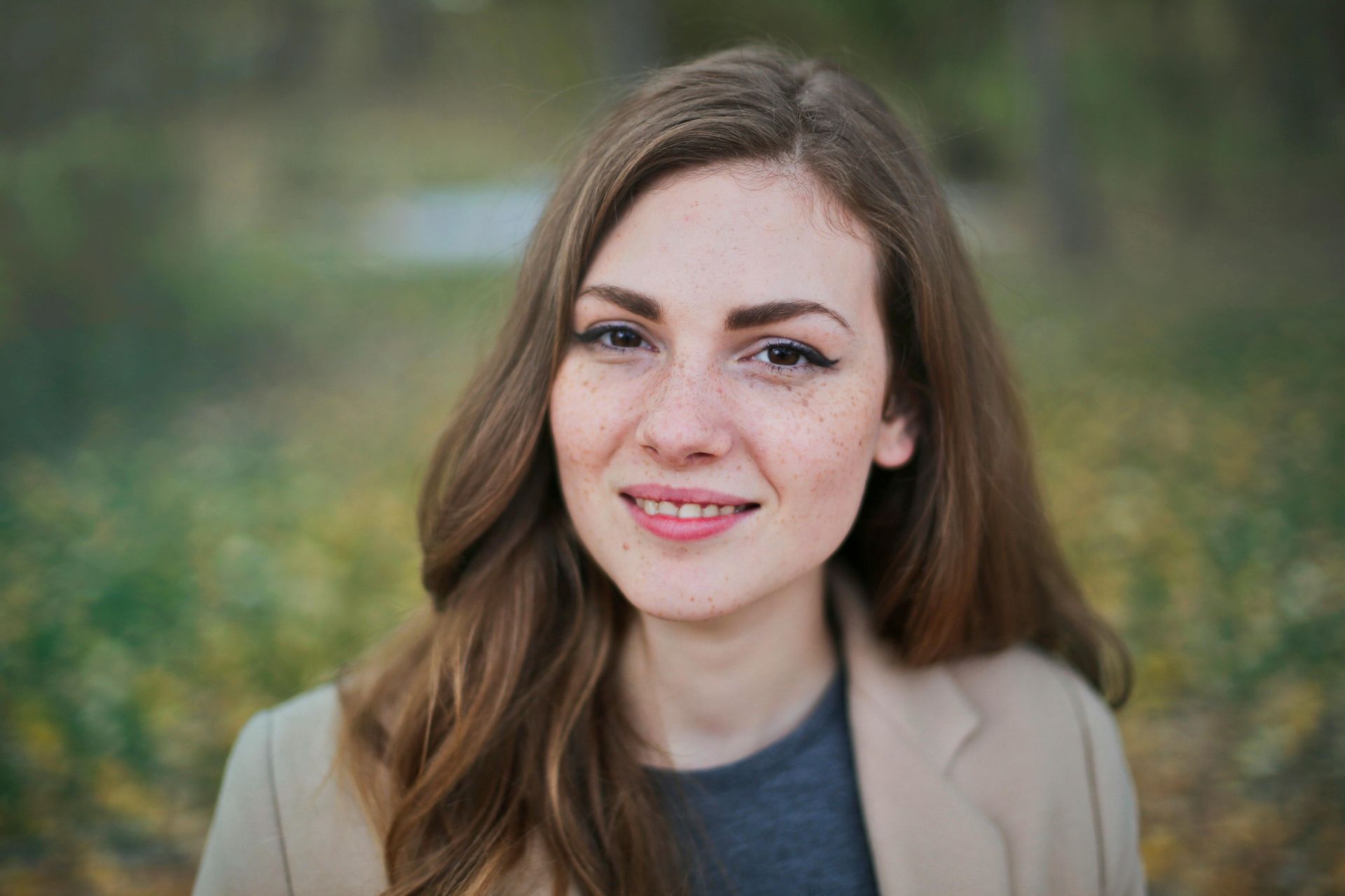 Woman with freckles smiles outdoors, wearing a tan blazer and gray shirt.