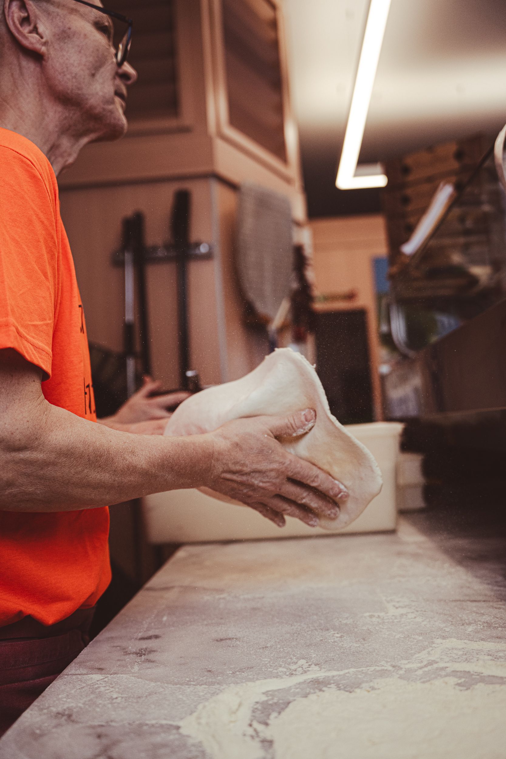 In una cucina, una persona con una maglietta arancione modella a mano l'impasto della pizza su un bancone infarinato.