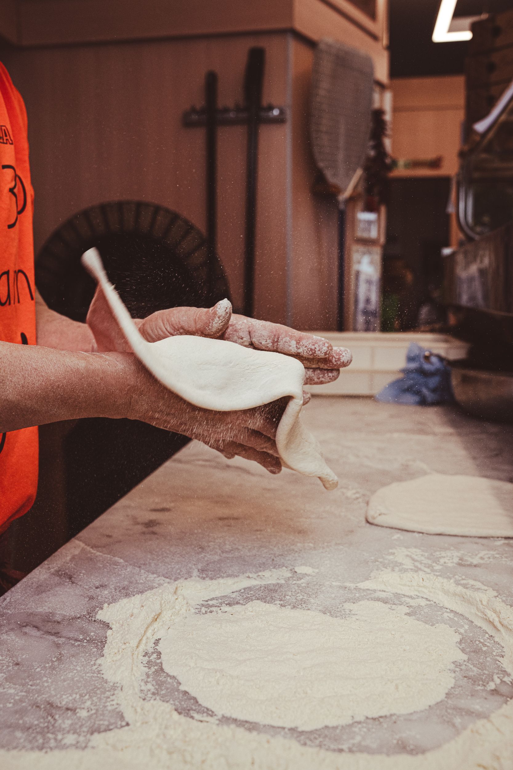 Una persona con una maglietta arancione stende l'impasto della pizza su un bancone della cucina cosparso di farina.