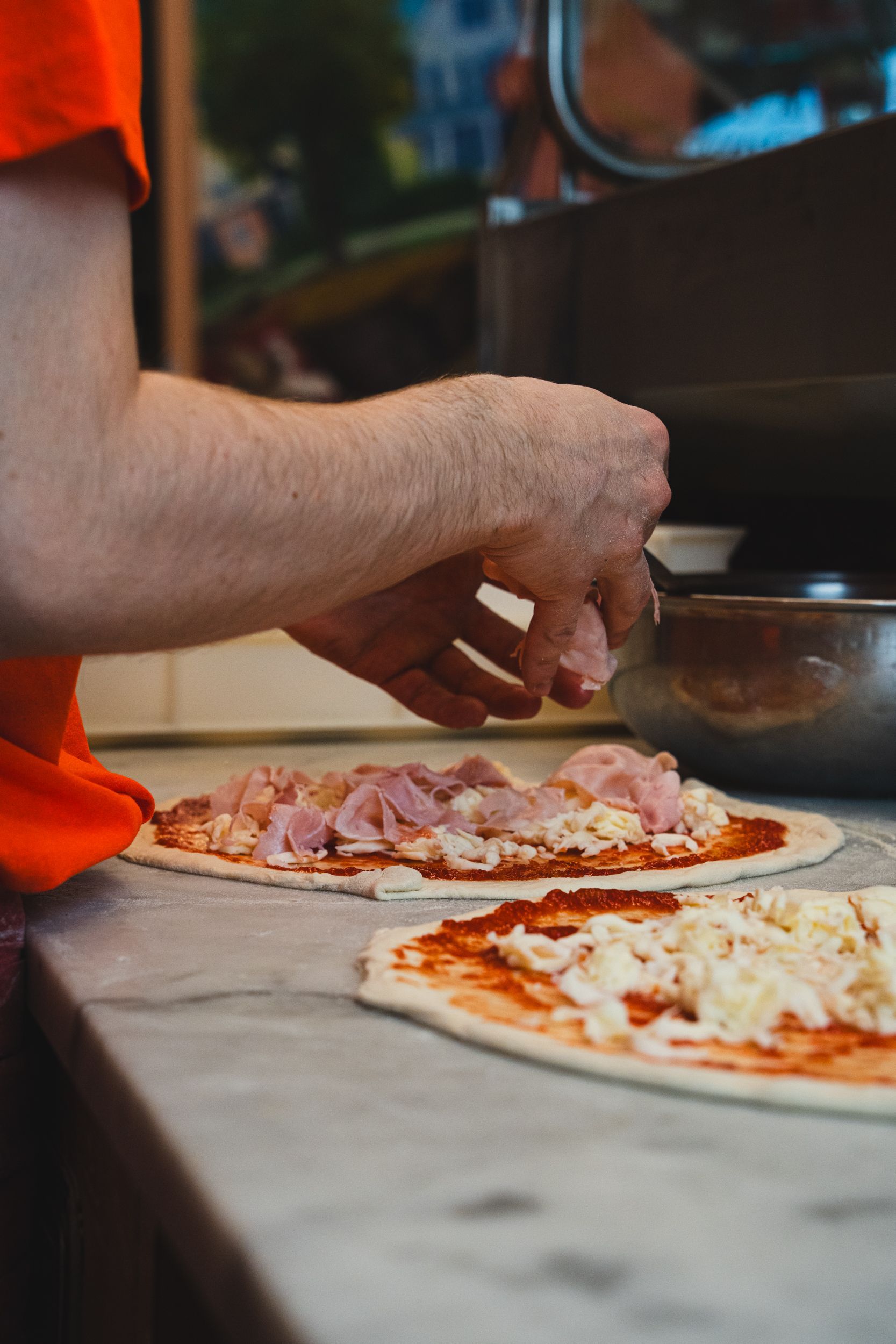 Una persona con una maglietta arancione aggiunge il prosciutto a una pizza su un piano di marmo in una cucina.