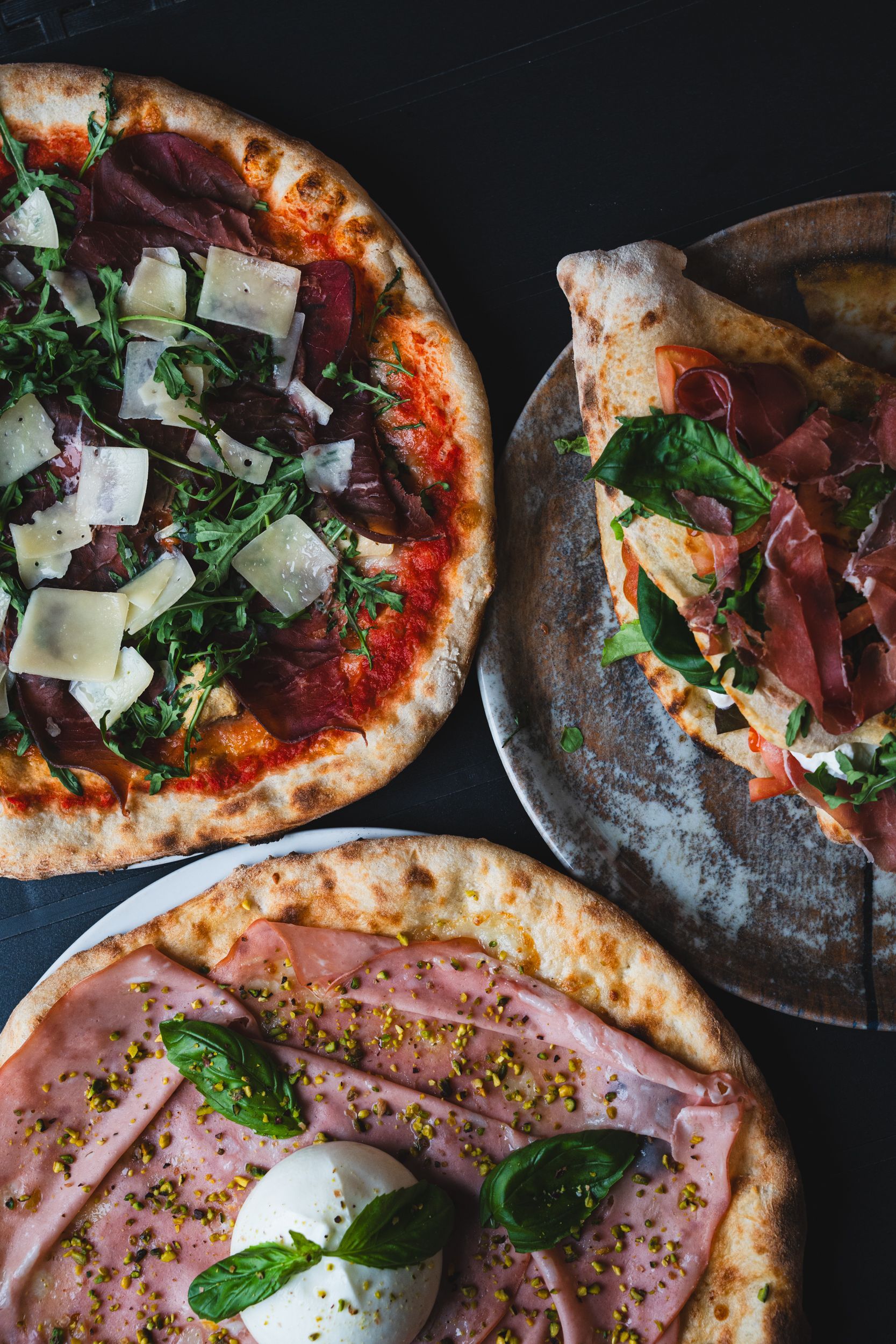 Tre pizze gourmet con vari condimenti, tra cui prosciutto, rucola, formaggio, mortadella e pistacchio, su un tavolo scuro.