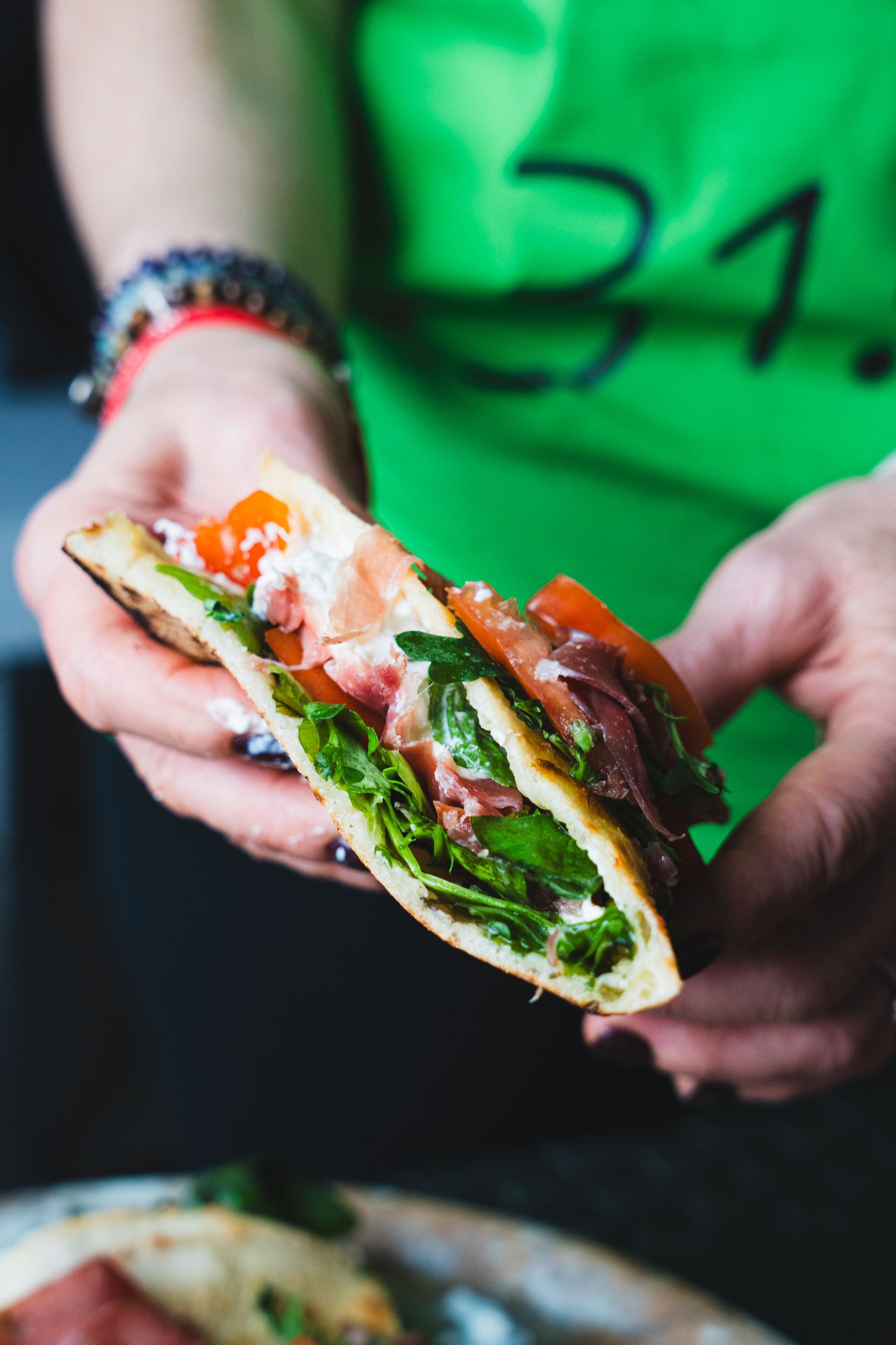 Primo piano di mani che reggono un panino con pane piatto piegato e farcito con verdure fresche, pomodori a fette e carne.