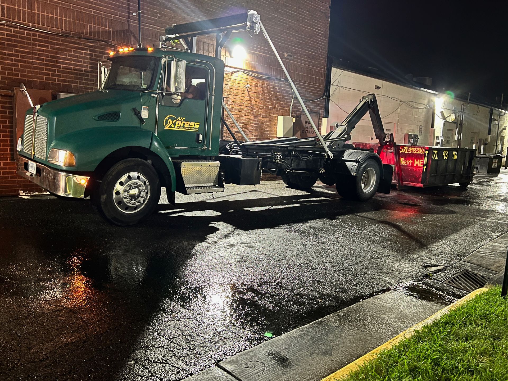 Green trash truck with a container in a wet alley at night.