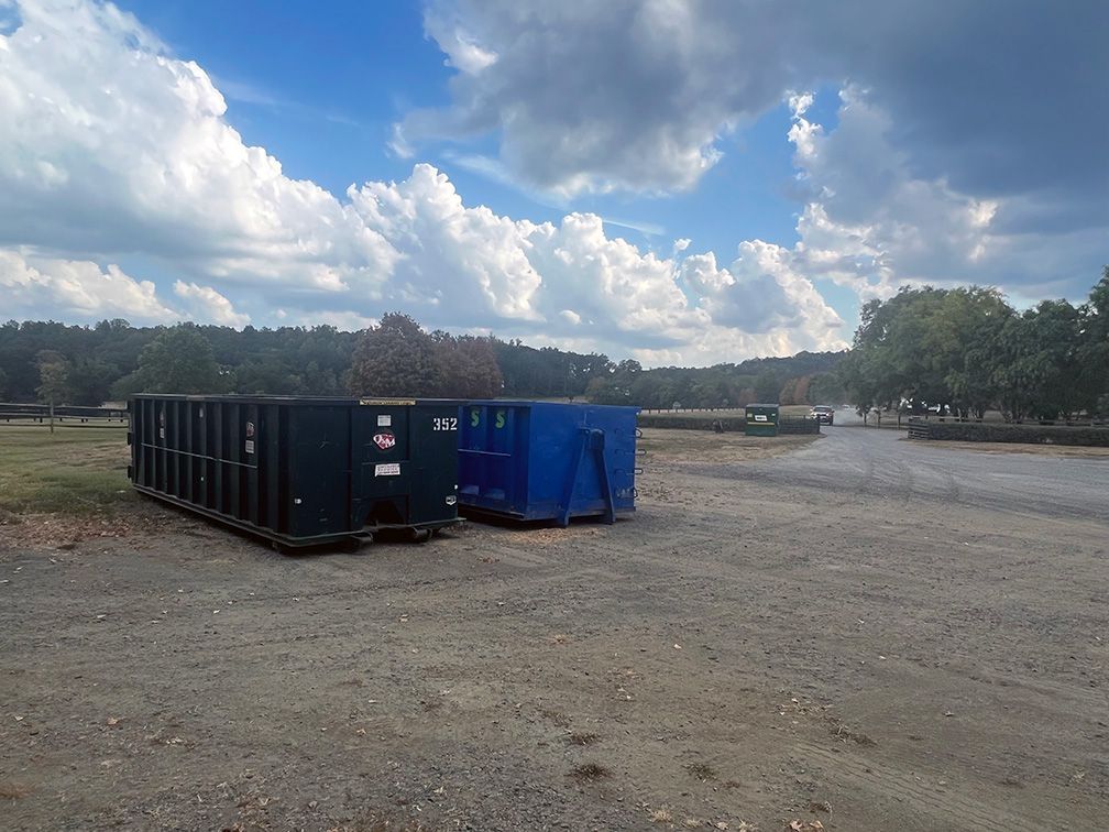 Two dumpsters are parked in a dirt lot with trees in the background.