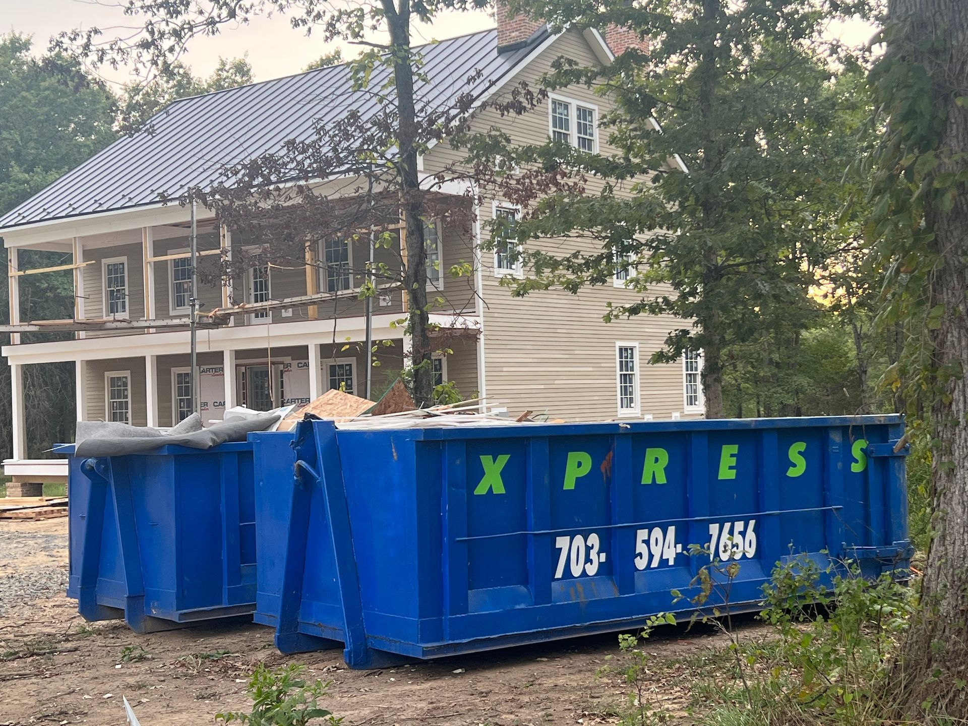 Blue dumpsters in front of a house under construction; trees and a phone number on the side.