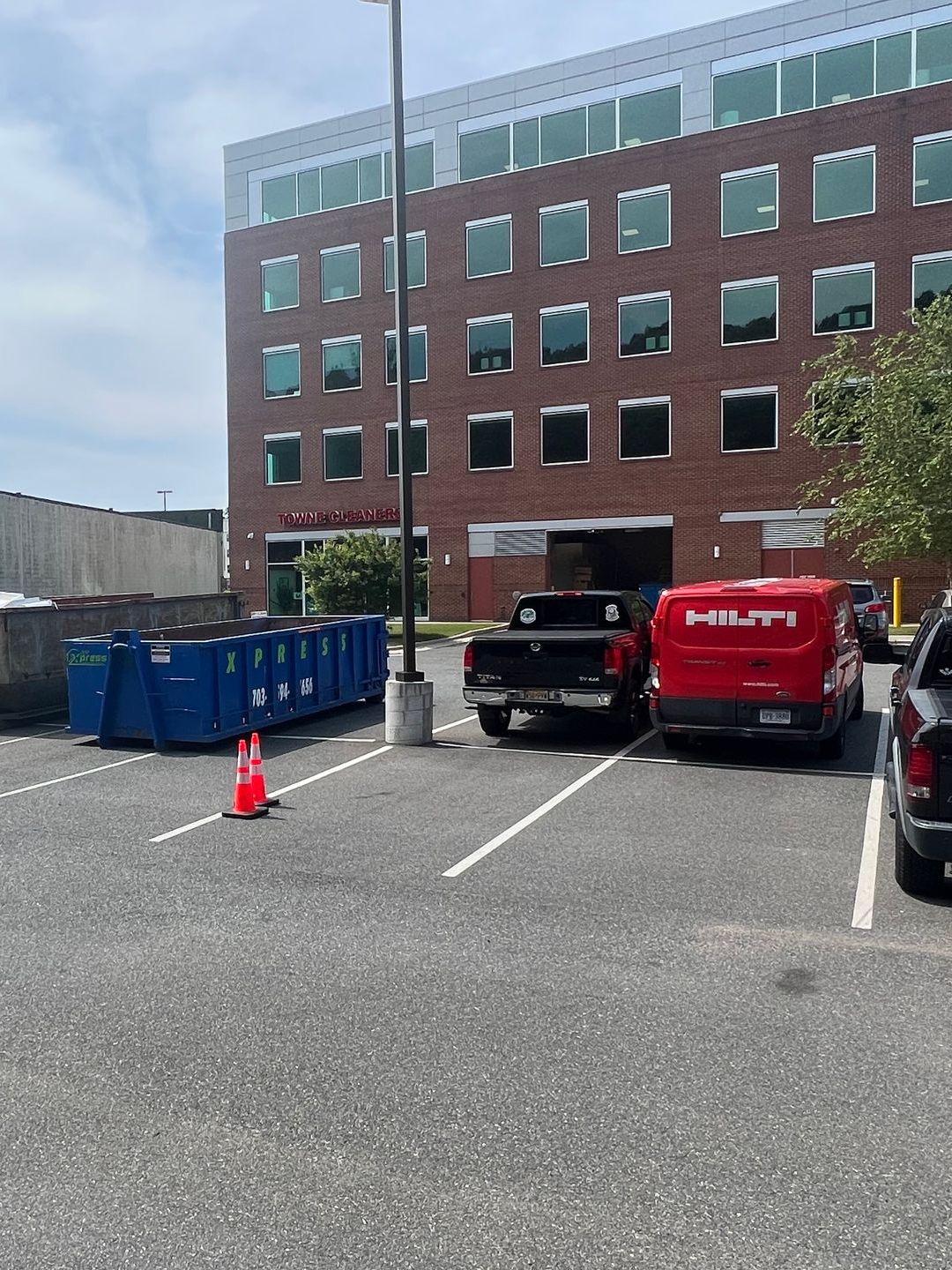 Parking lot with a brick building. Dumpsters, work trucks, and a traffic cone are present.