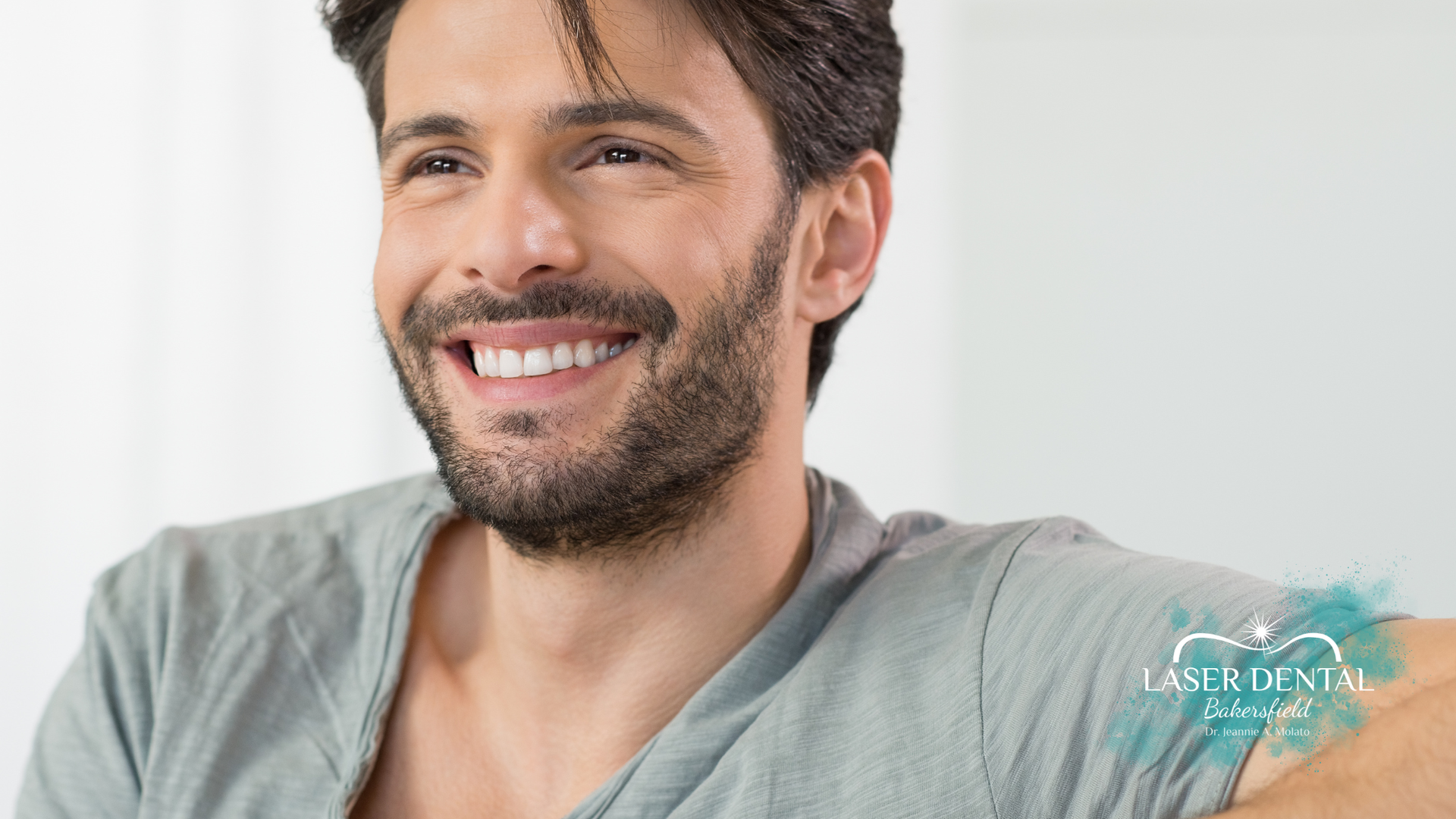 Man smiling with bright white teeth, wearing a gray shirt, against a blurred background.