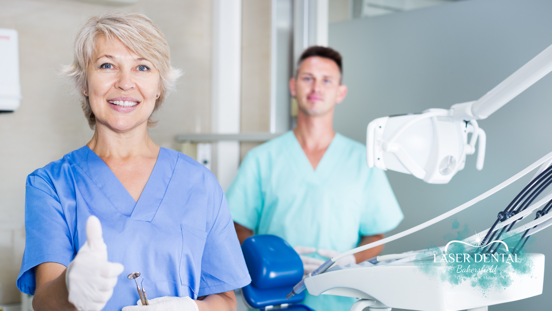 Dentist and assistant smiling, in a dental office. The dentist is giving a thumbs up.