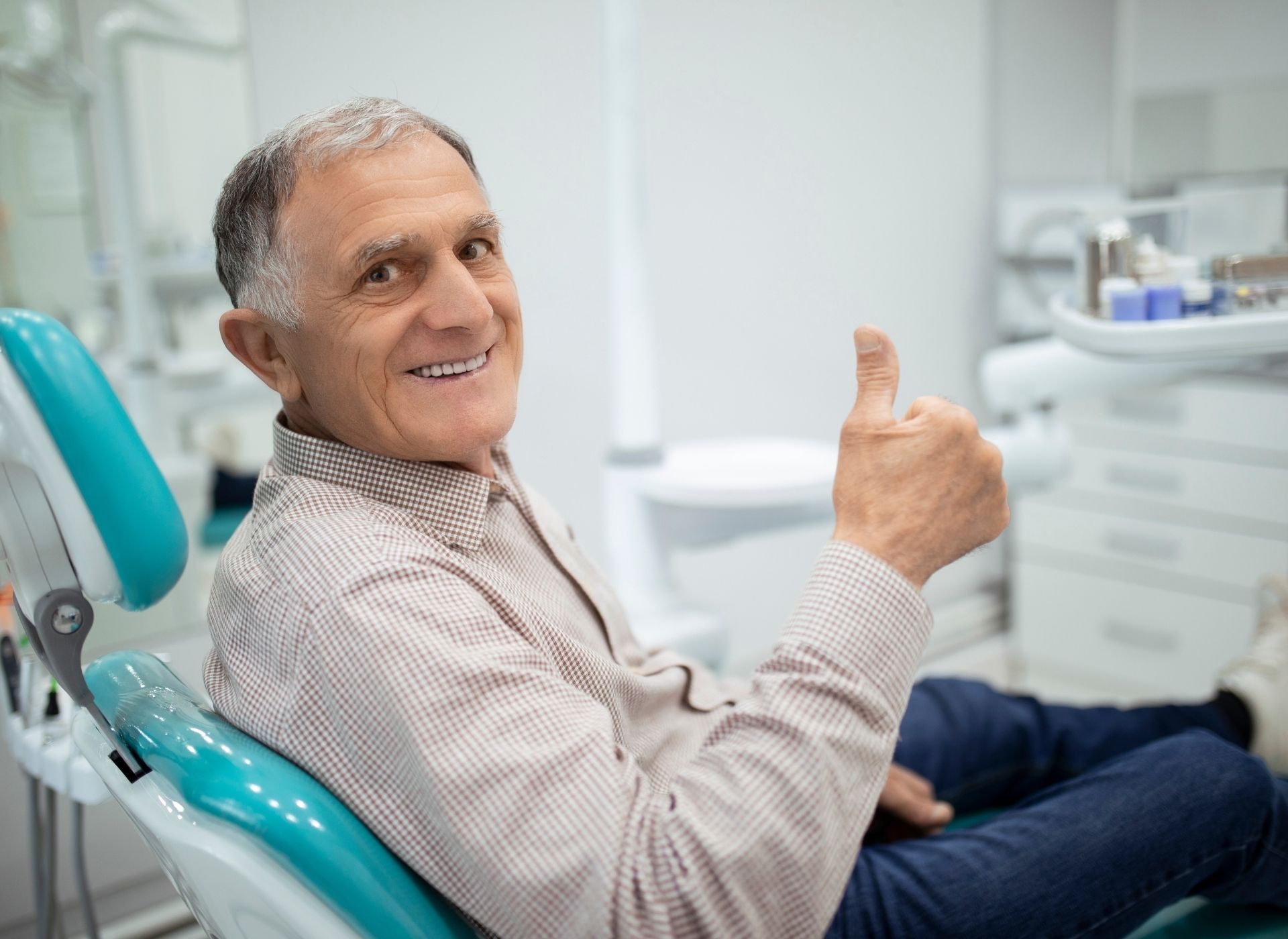 Man in dental chair gives thumbs up, smiling. Tan shirt, blue jeans. Dentist's office setting.
