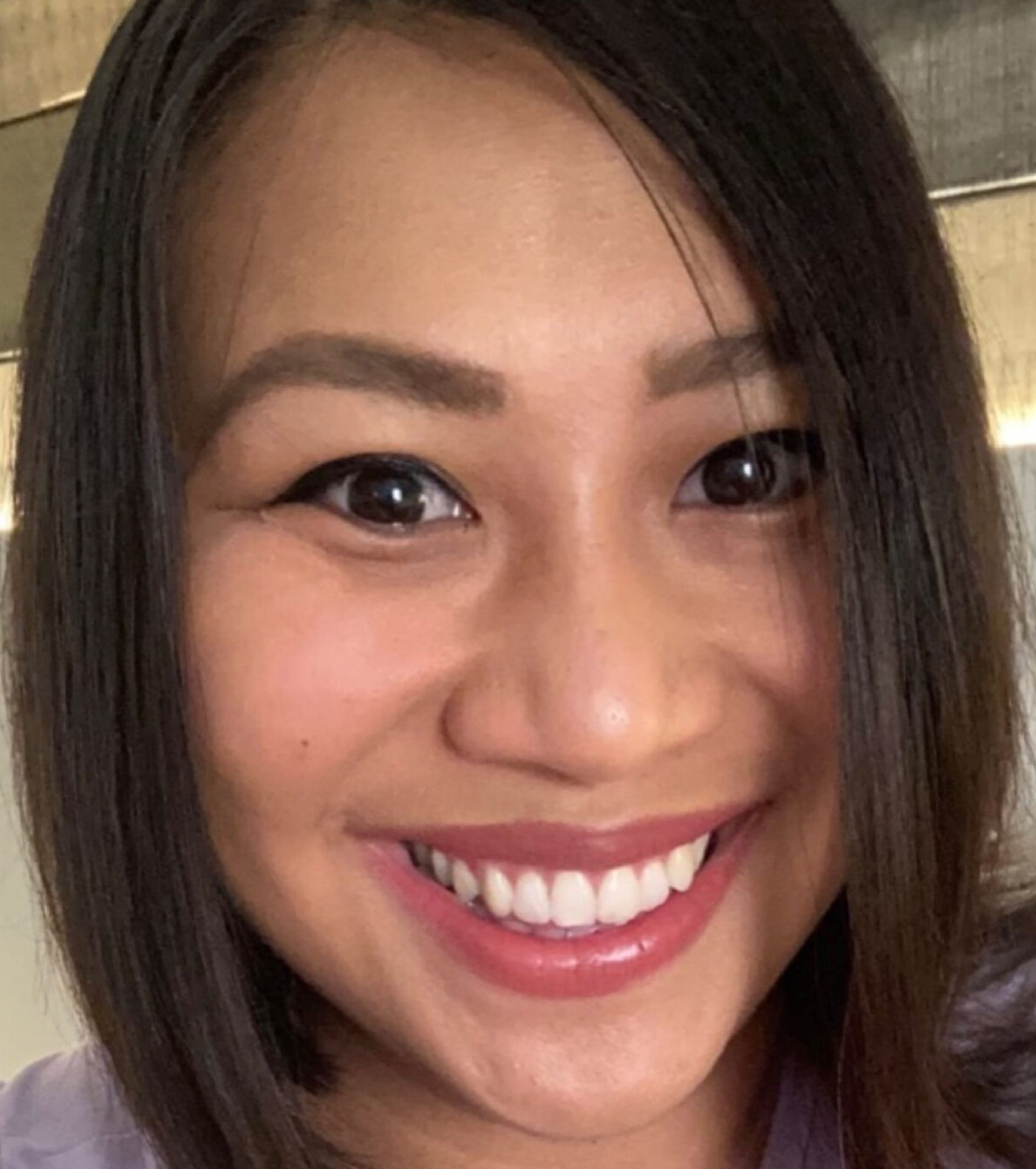 Woman with dark hair smiles widely, showing white teeth. She has brown eyes and wears a purple shirt.