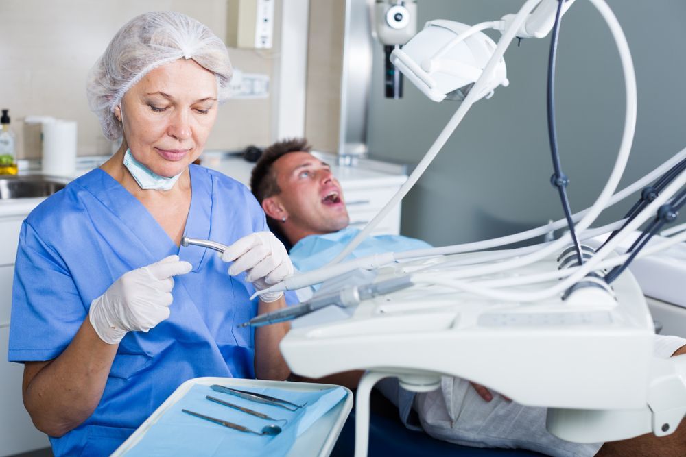 Dentist in blue scrubs examines a male patient's teeth with instruments in a dental office.