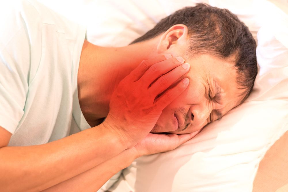 Man in bed, clutching his jaw in pain, red highlighting jaw and neck.
