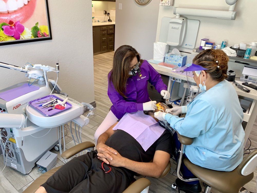 Dental procedure: Patient in chair, dentist in purple and assistant in blue working on patient's mouth.