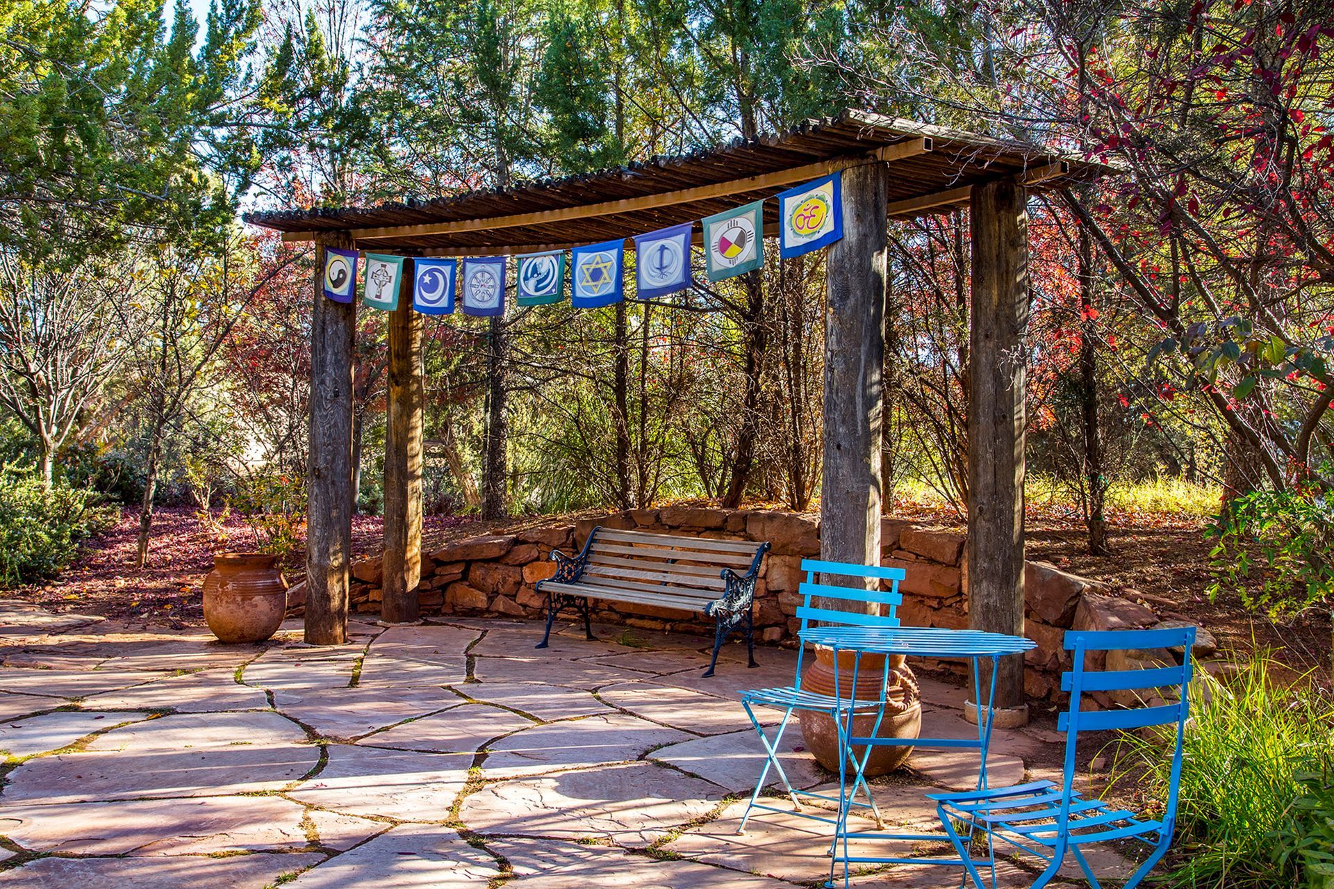 There is a bench and chairs under a pergola in the woods.