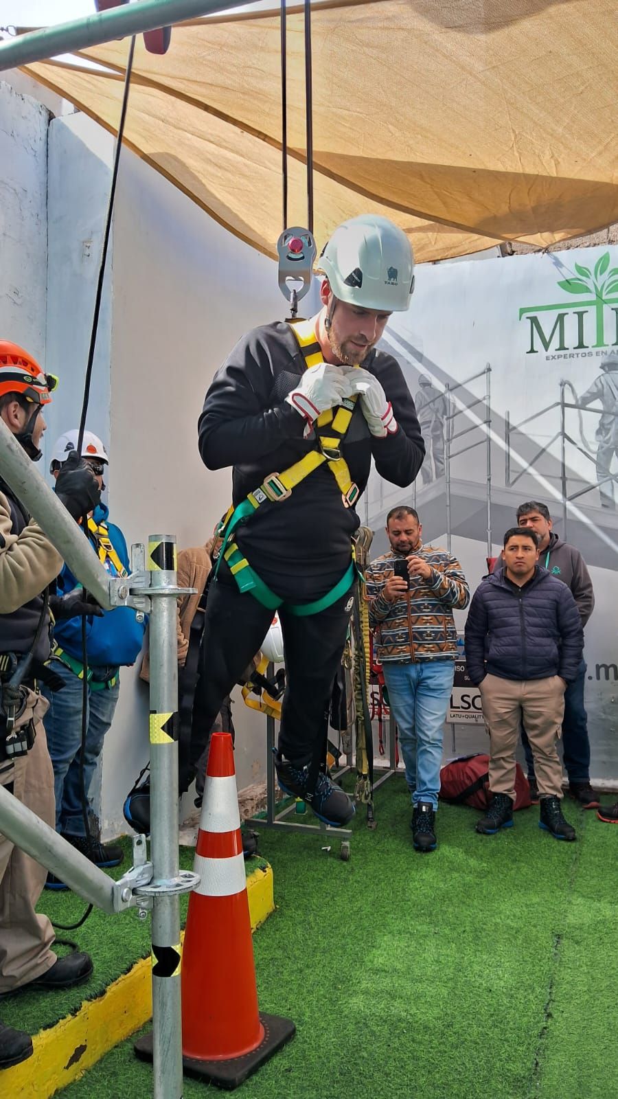 Hombre con arnés de seguridad suspendido, practicando un rescate desde un andamio, observado por un grupo.