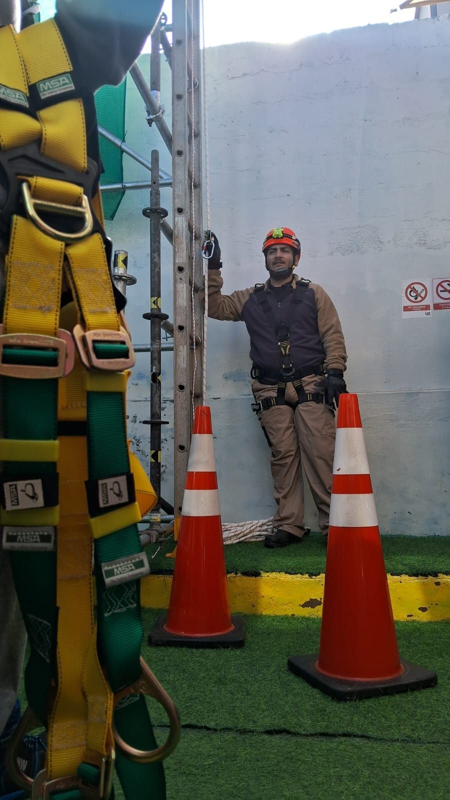 Trabajador con equipo de seguridad junto a escalera y conos, contra pared blanca.