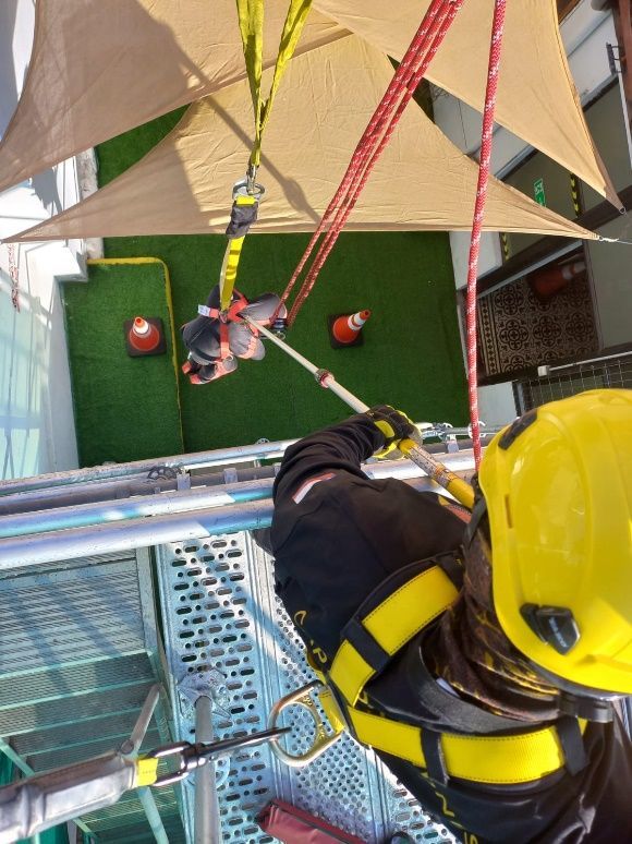 Persona con casco amarillo y arnés haciendo rapel desde una altura, con cuerdas de seguridad.