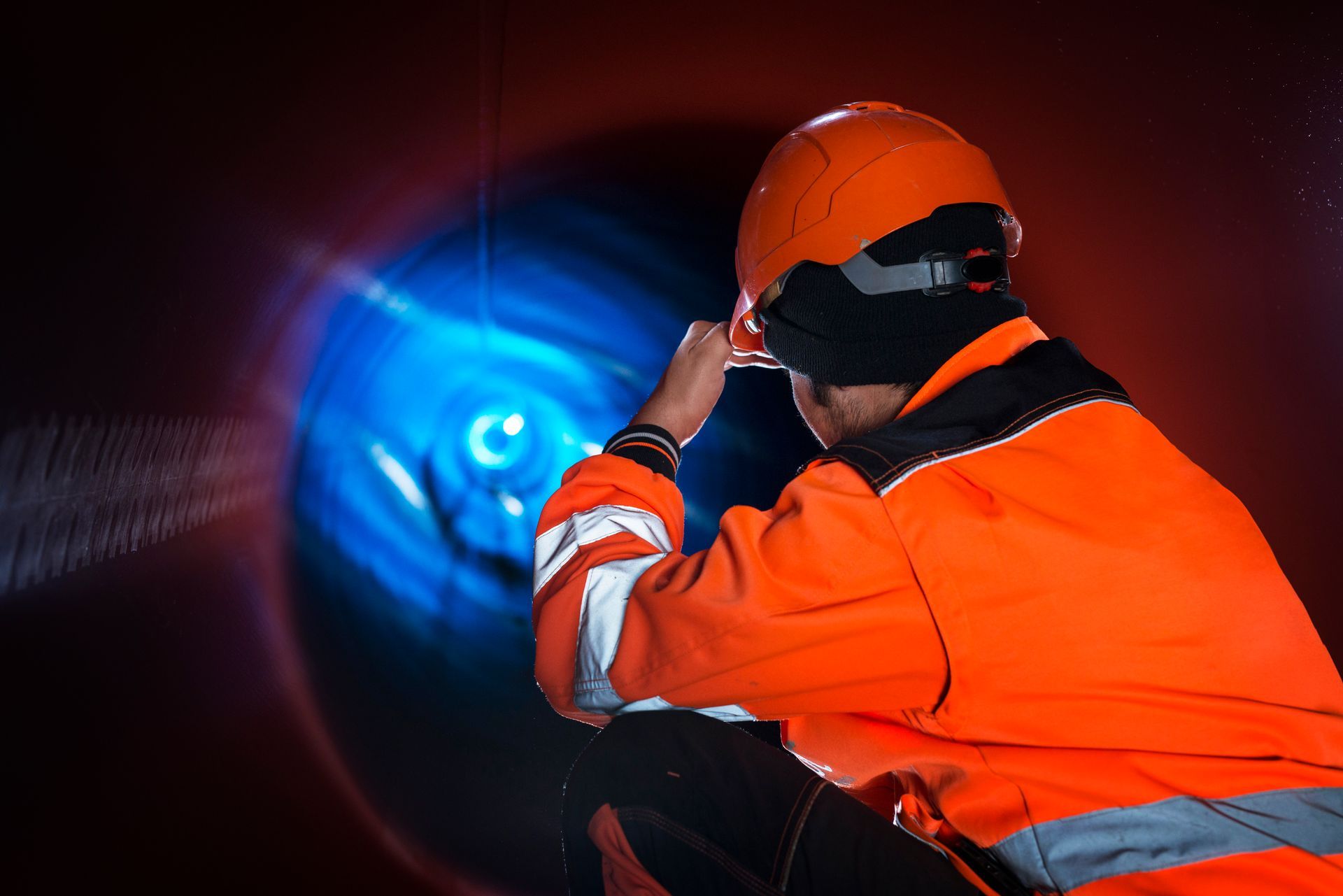 Trabajador con equipo de seguridad naranja inspeccionando un túnel iluminado de azul, usando un casco y sosteniendo una linterna.