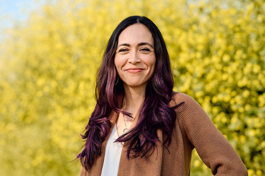 A woman with purple hair is standing in front of a field of yellow flowers.