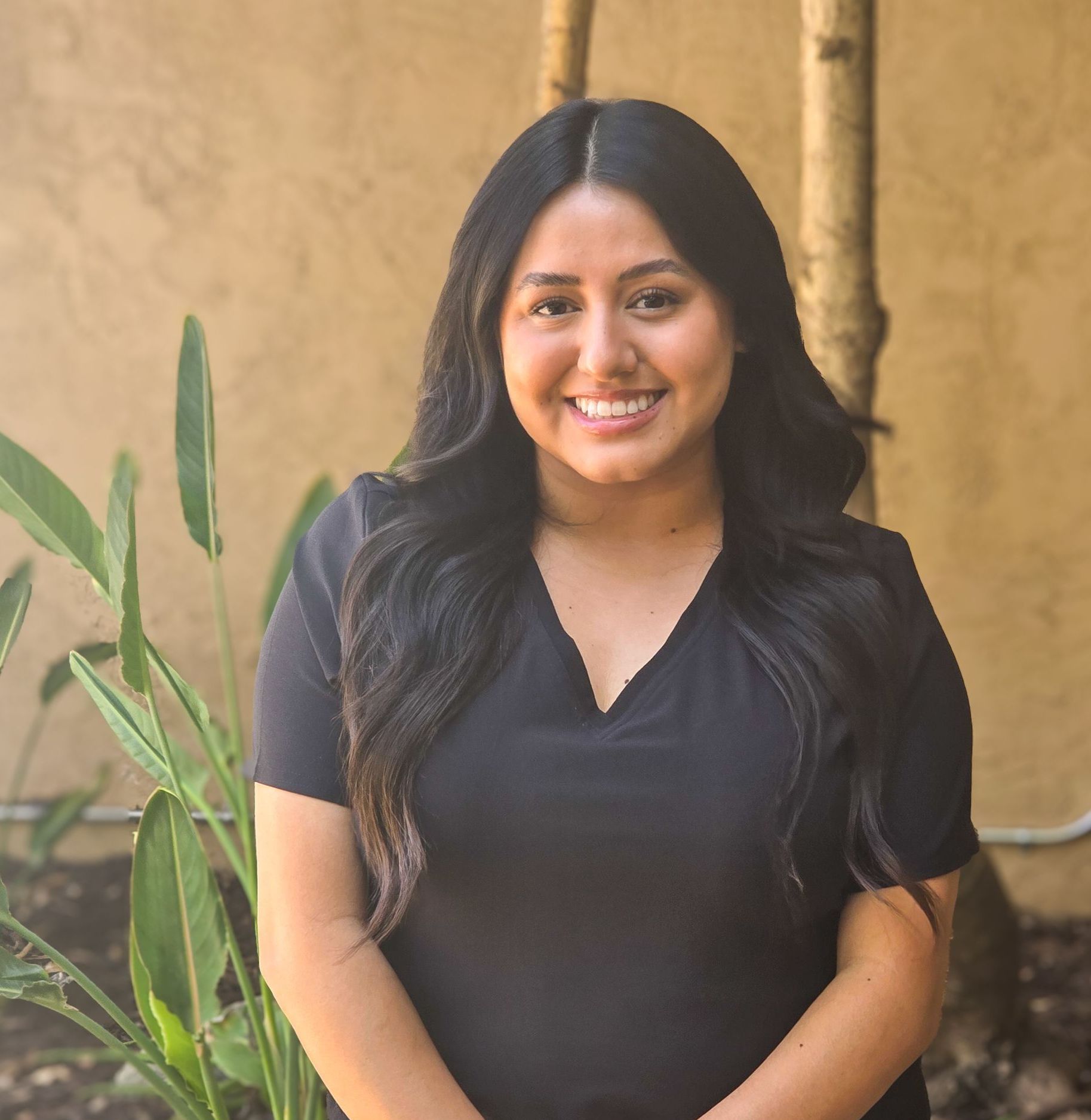 Woman with long dark hair smiles, standing near a tree. Wearing a black v-neck shirt.