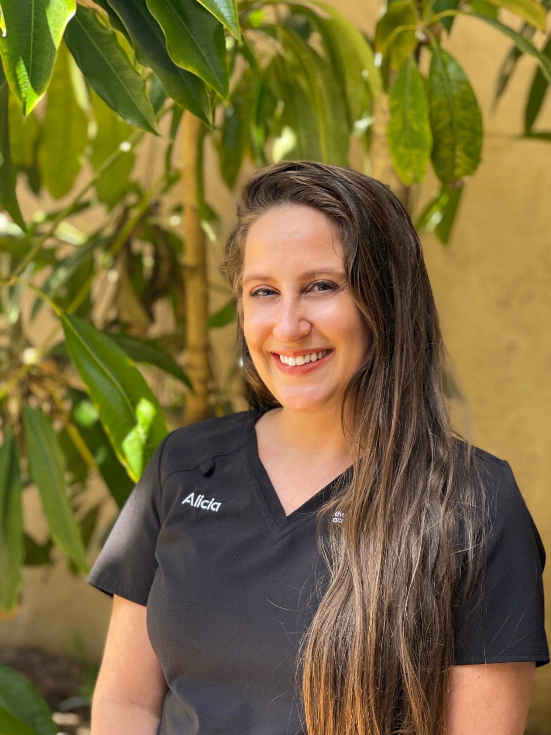 Woman with long brown hair smiling, wearing a black uniform, outdoors near green leaves.