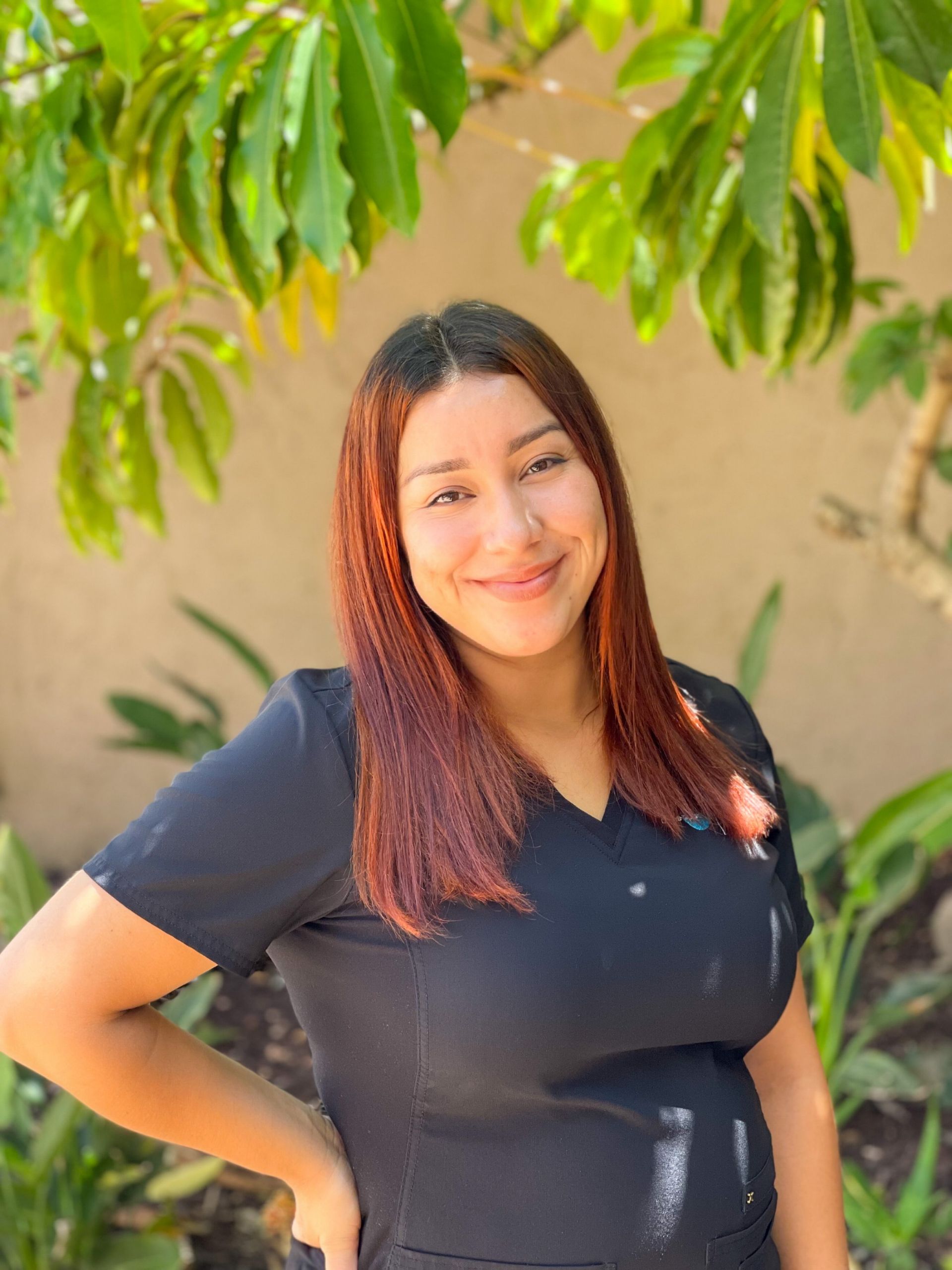 Woman with auburn hair smiles outdoors, wearing black scrubs, posing near greenery.