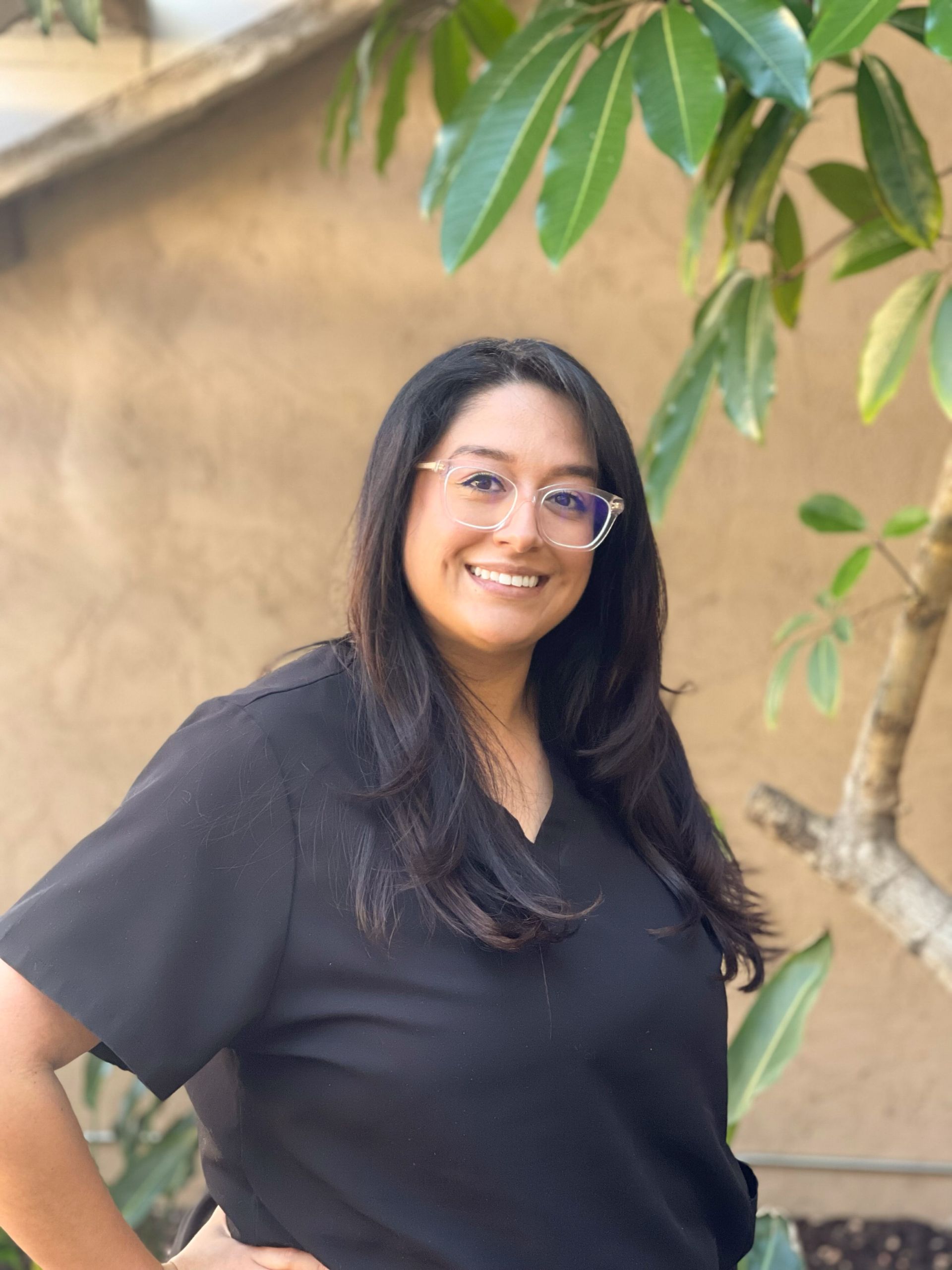 Woman with glasses smiles, wearing black scrubs, standing outdoors near green leaves.