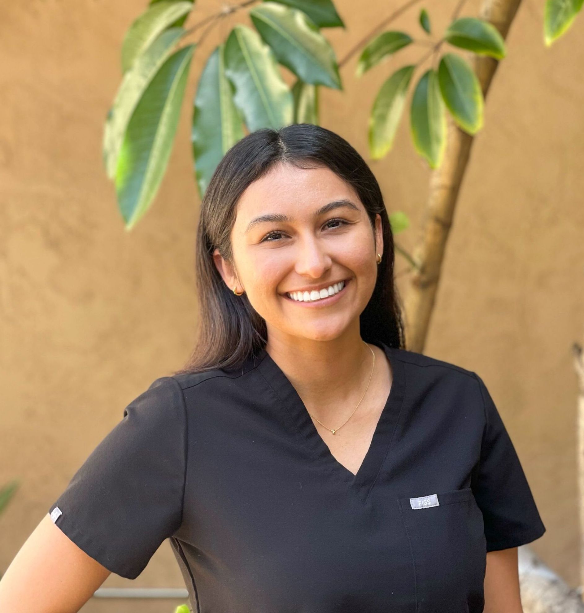 Woman in black scrubs smiling, leaning against a tree, outdoors.